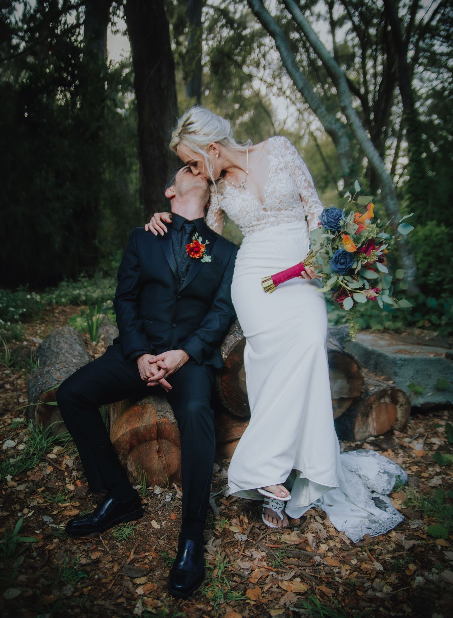 A bride and groom share a romantic kiss outdoors, she in a lace gown holding a colorful bouquet, he in a black tuxedo.