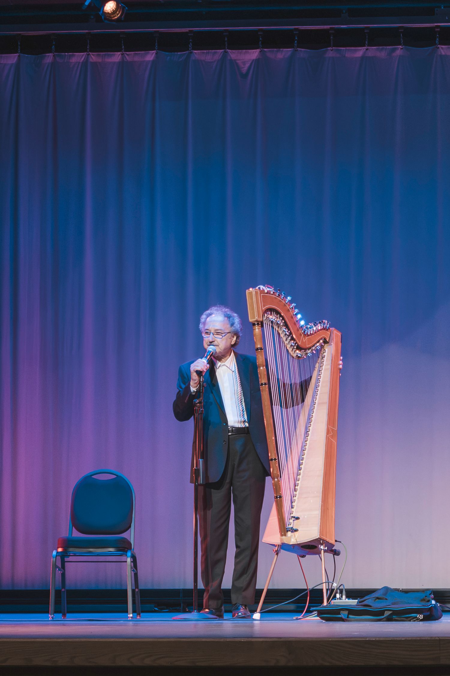 Man speaking on stage with a harp and empty chair under blue and purple stage lighting.