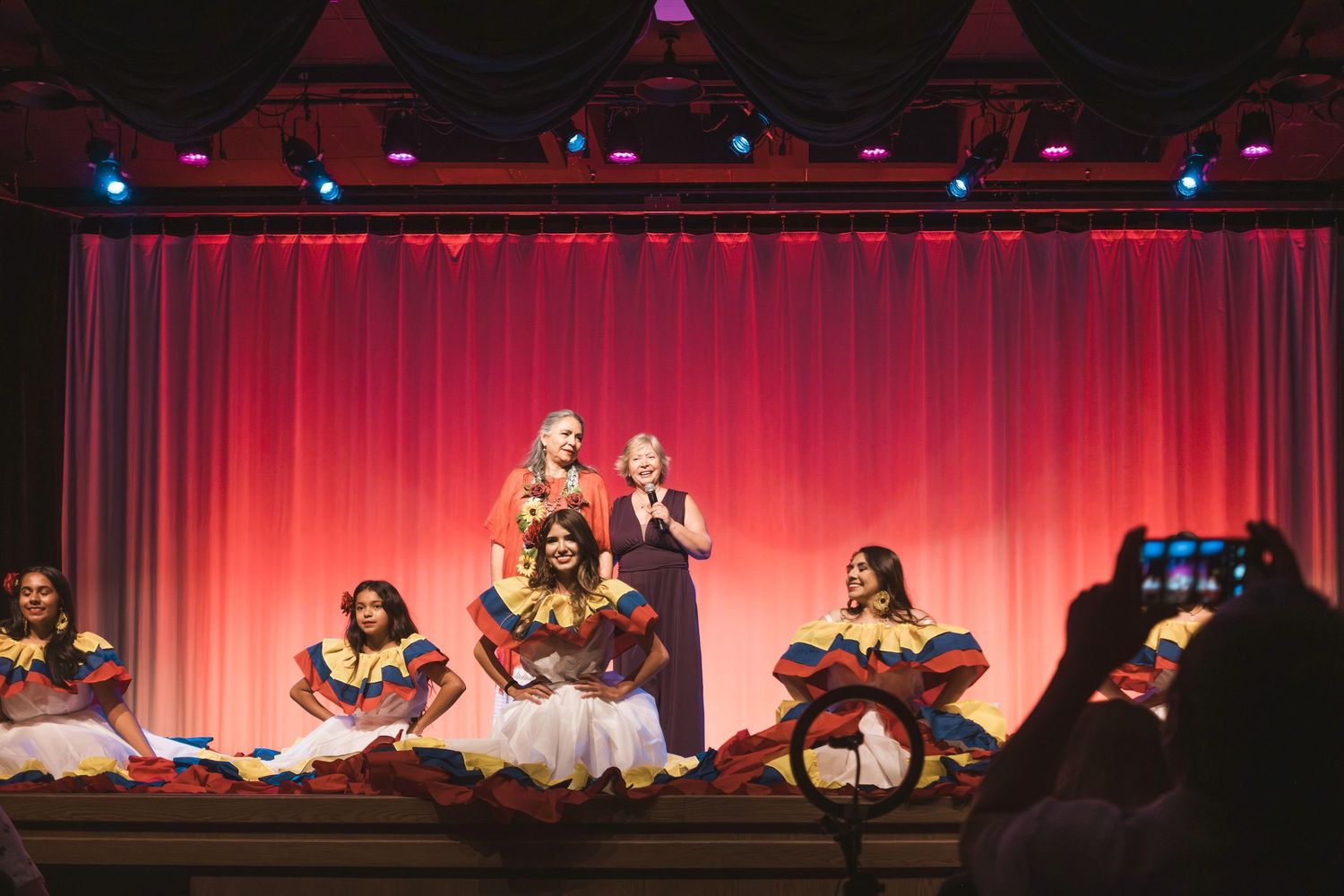 Performers in colorful Colombian dresses on a red-curtained stage with two women in blue gowns at center.