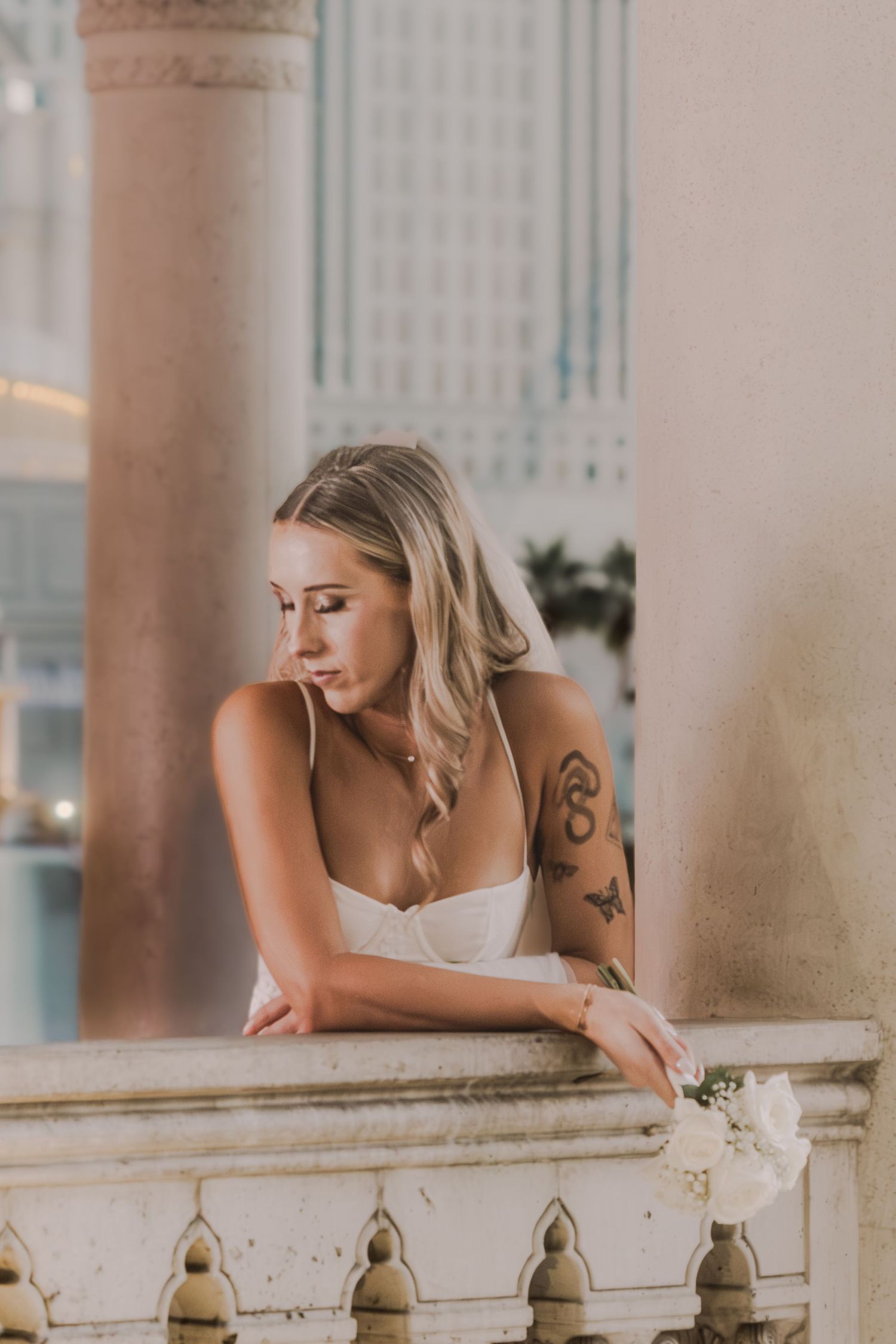 Blonde bride in white gown with floral tattoos leans on ornate balcony railing holding white flowers.