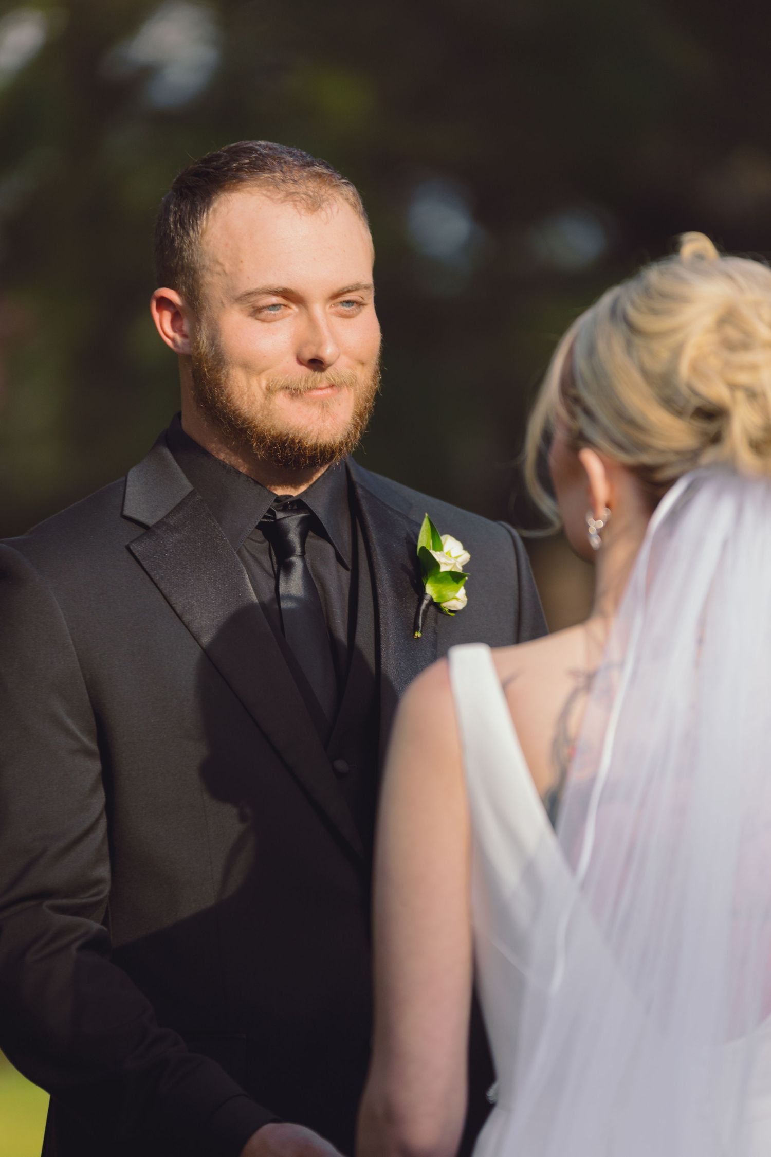 Bearded groom in black suit with boutonniere exchanging vows with bride outdoors.