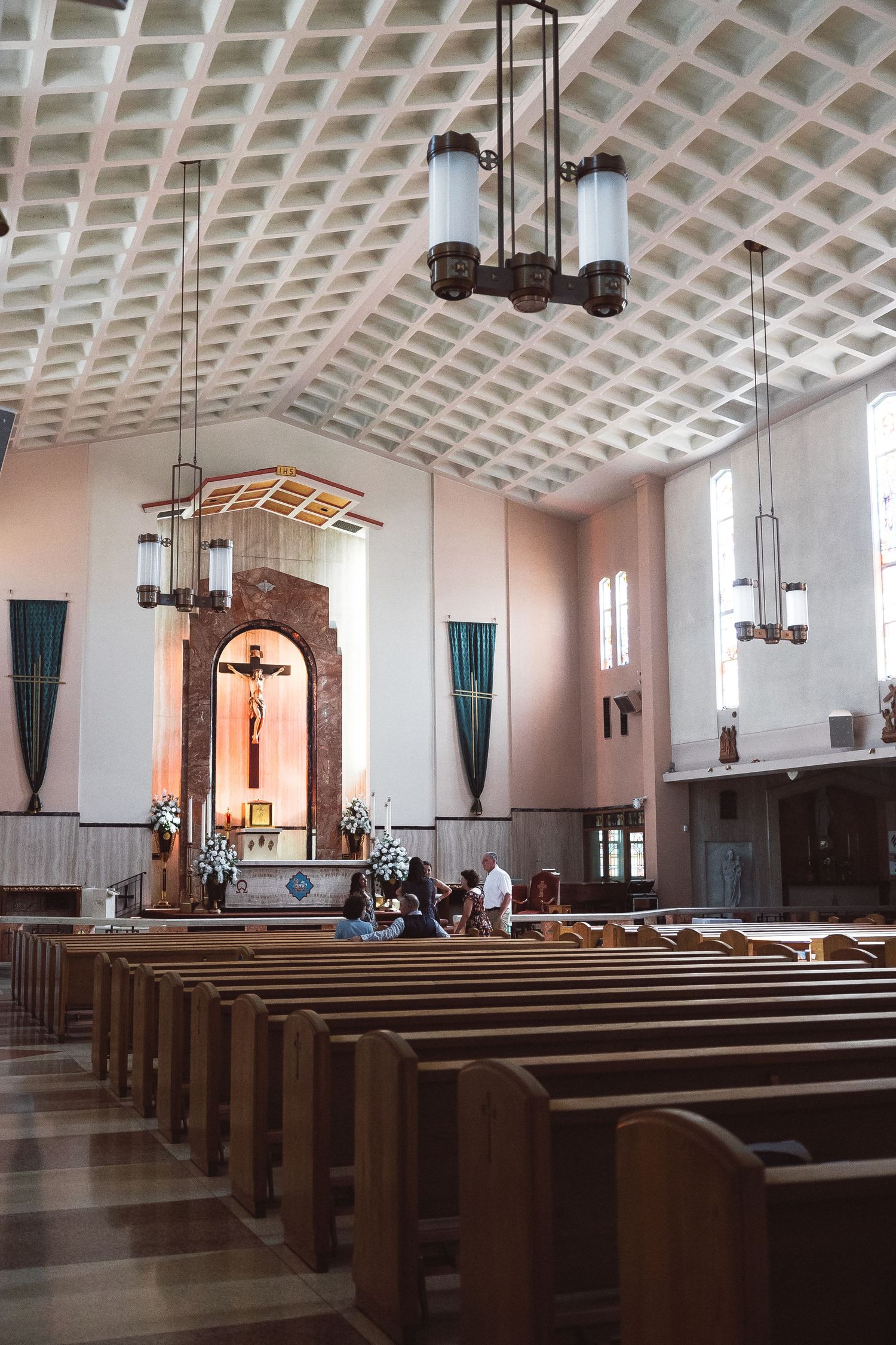 Catholic church interior with wooden pews, crucifix altar, coffered ceiling, and elegant pendant lights.