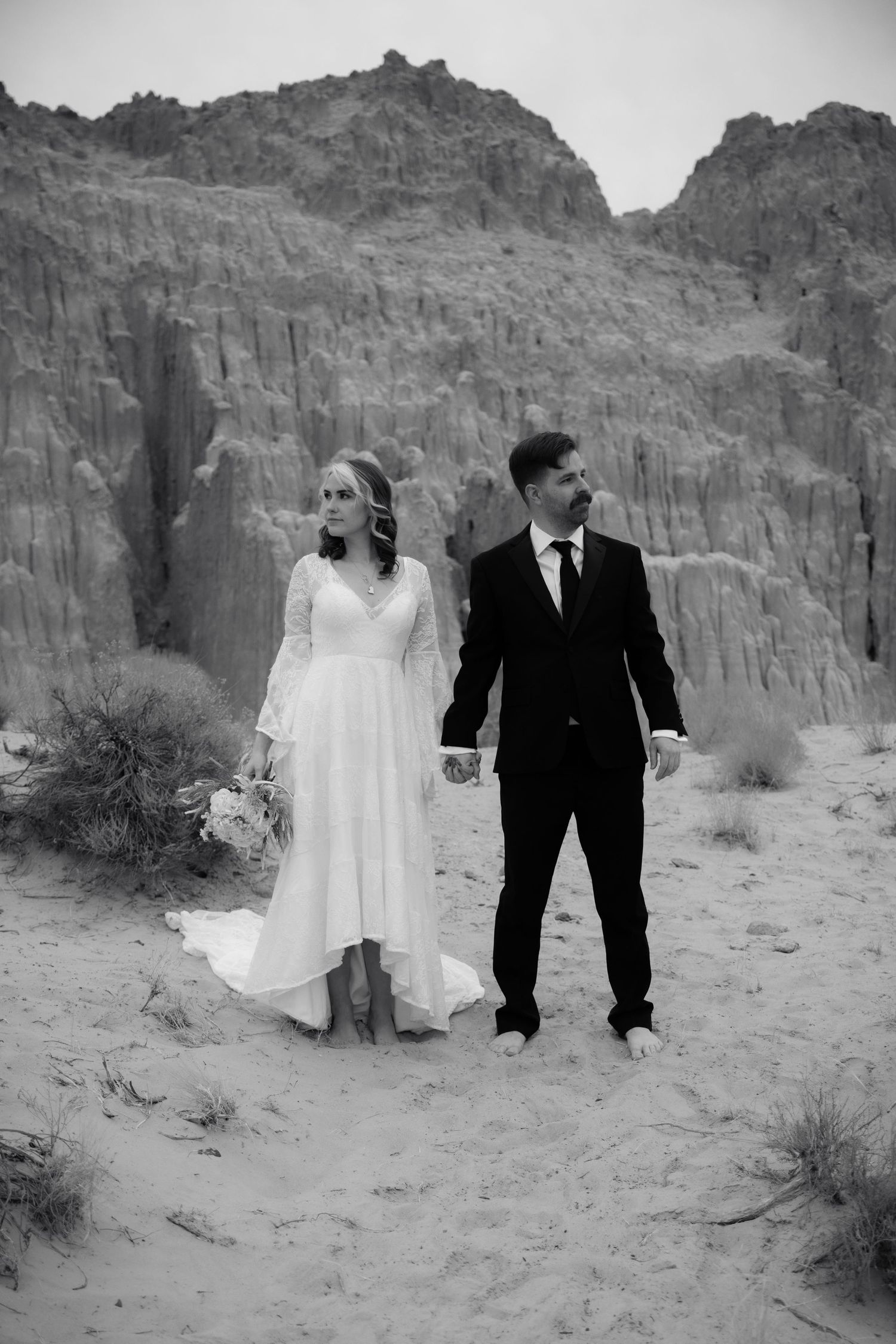 Black and white photo of barefoot bride and groom holding hands in desert with towering rock columns.