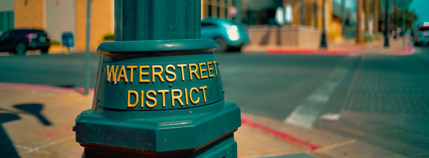 Green Waterstreet District sign post on a city street corner with teal buildings and parked cars in the background.