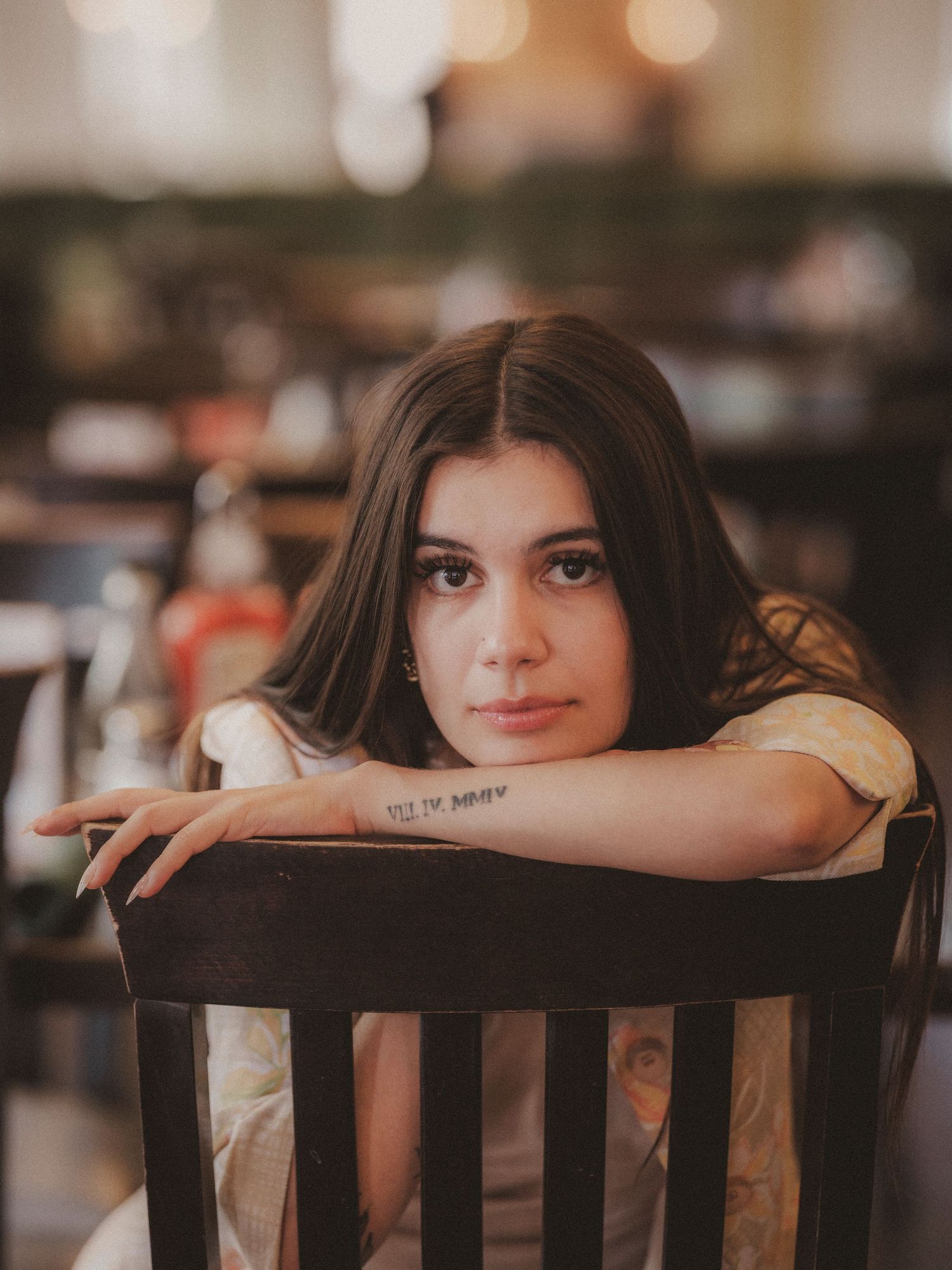 A young brunette woman with a forearm tattoo rests her chin on her arms over a dark chair.