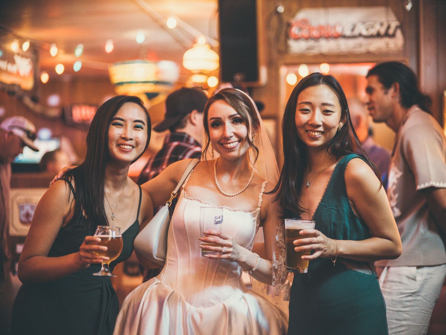 Bride and two bridesmaids smiling and holding drinks at a lively wedding reception party.