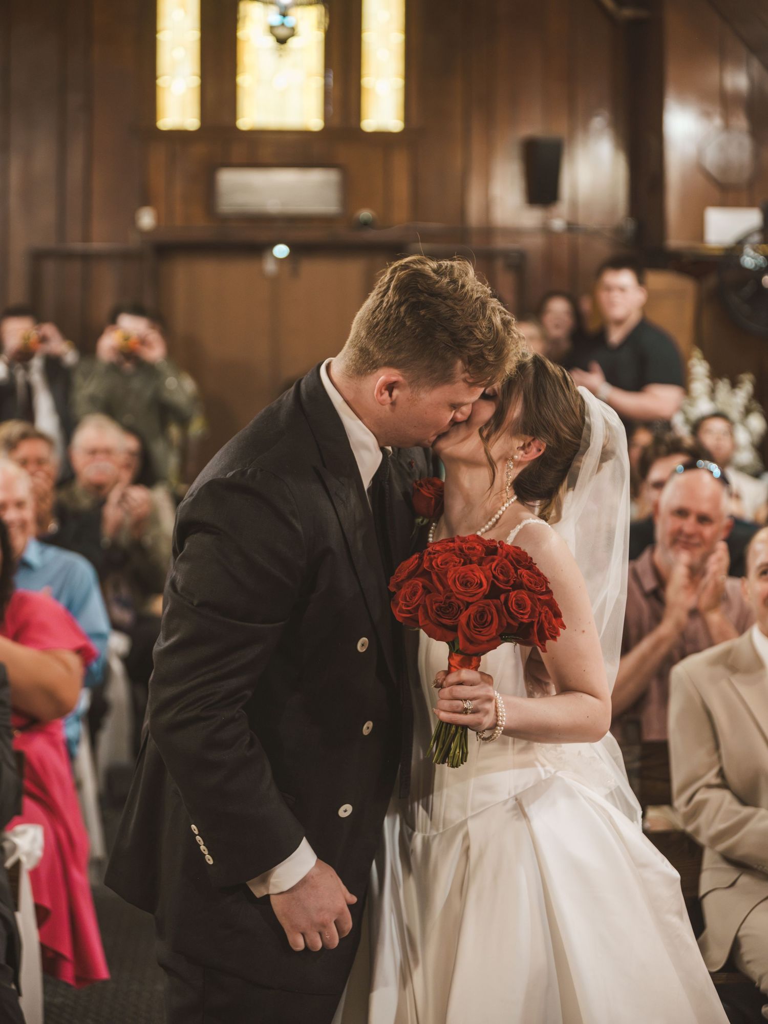 Newlywed couple sharing first kiss at wedding ceremony altar, bride holding red roses bouquet, guests applauding in church.