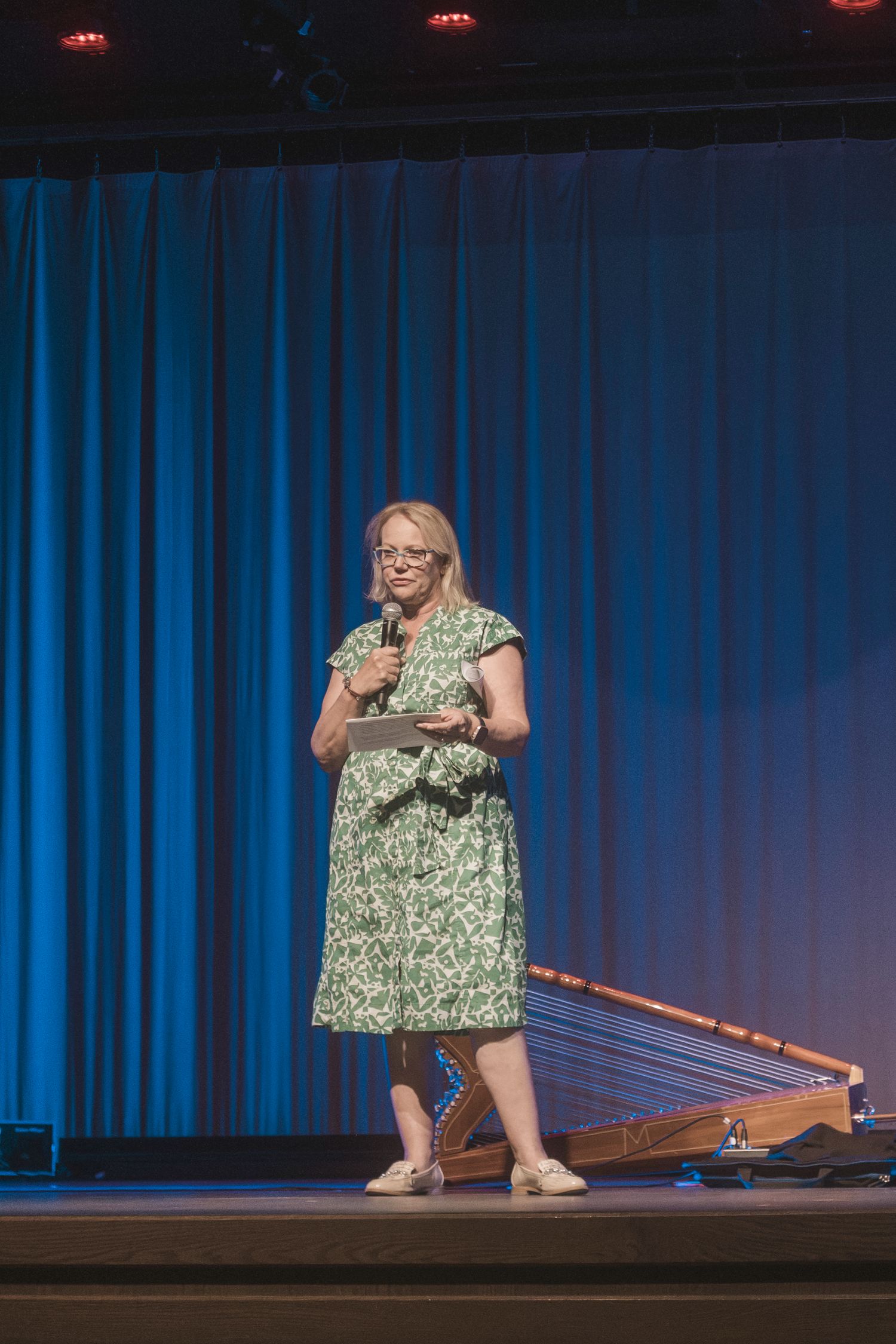 Woman in green floral dress speaks on stage holding microphone and tablet at a conference event.