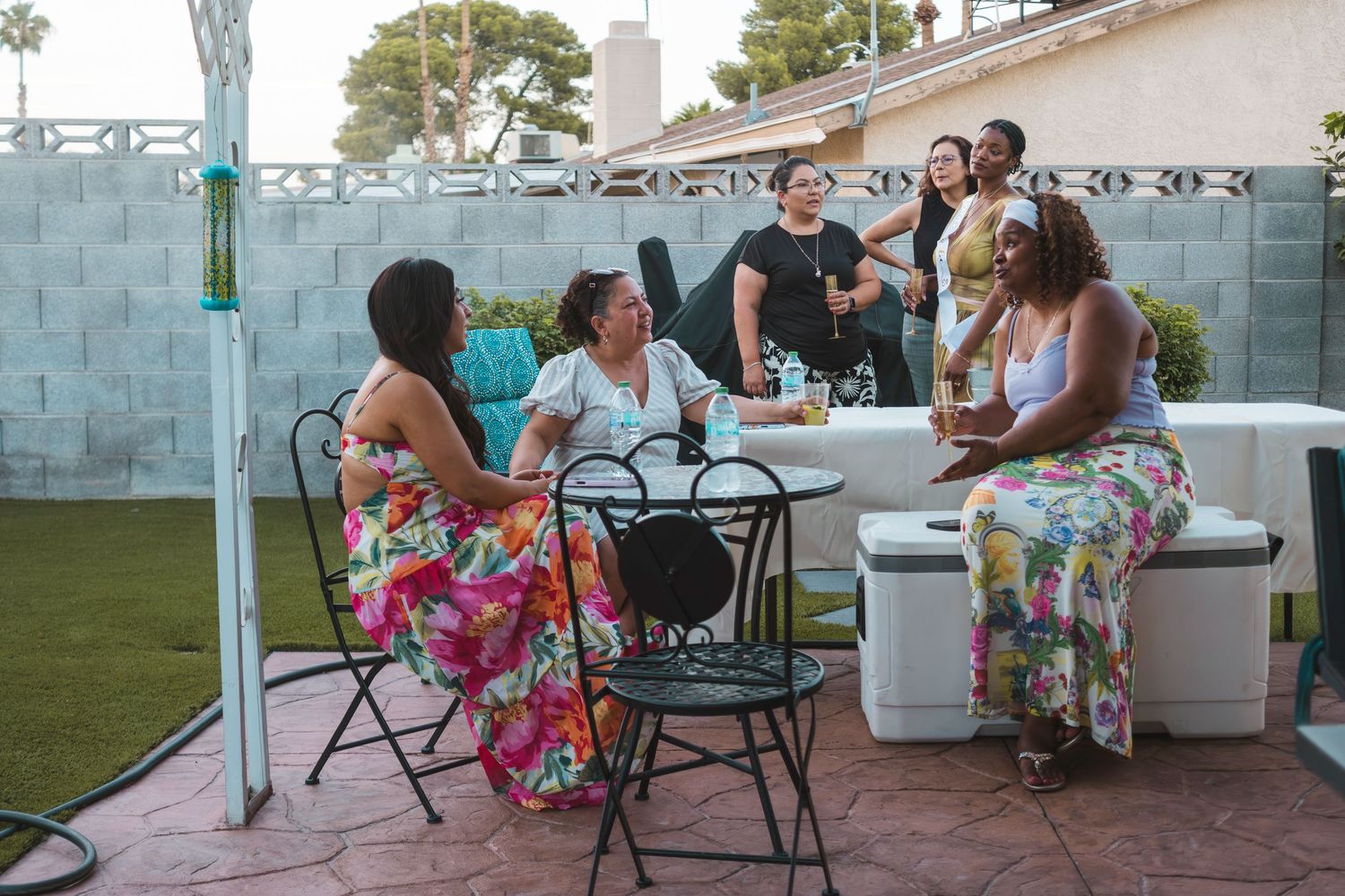 Women socializing outdoors at a backyard gathering, wearing colorful floral outfits, seated around a table on a patio.