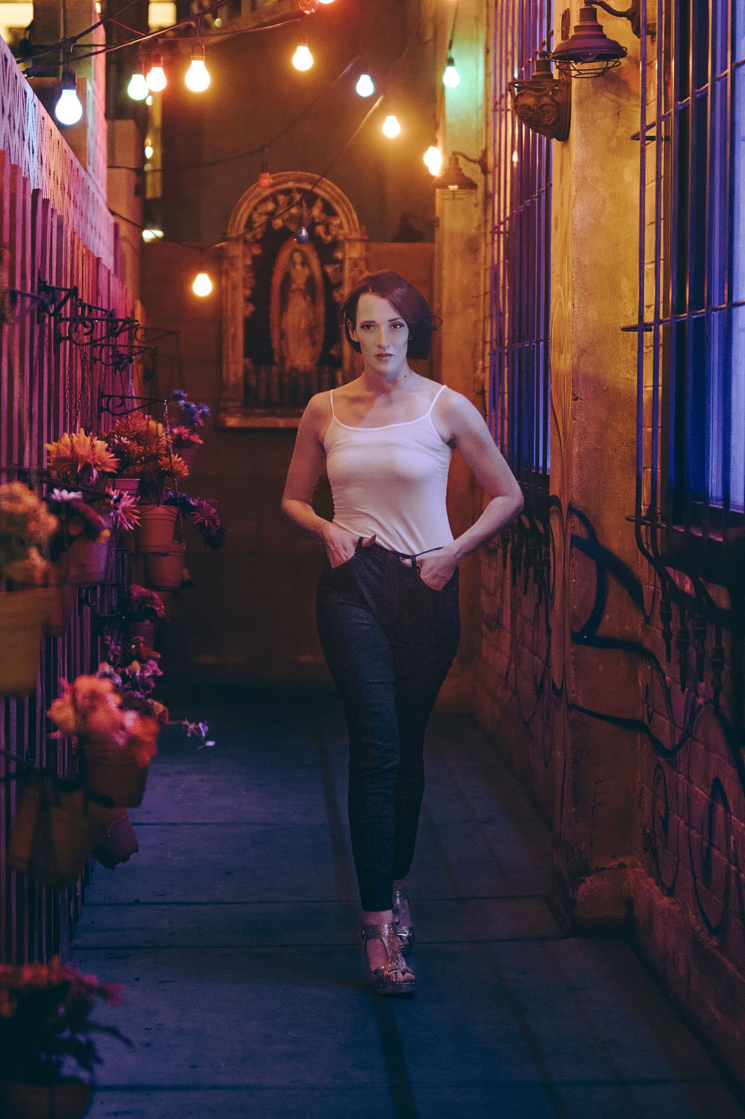 Woman in white tank top and black jeans poses in a narrow alley lit by string lights and colorful flowers at night.