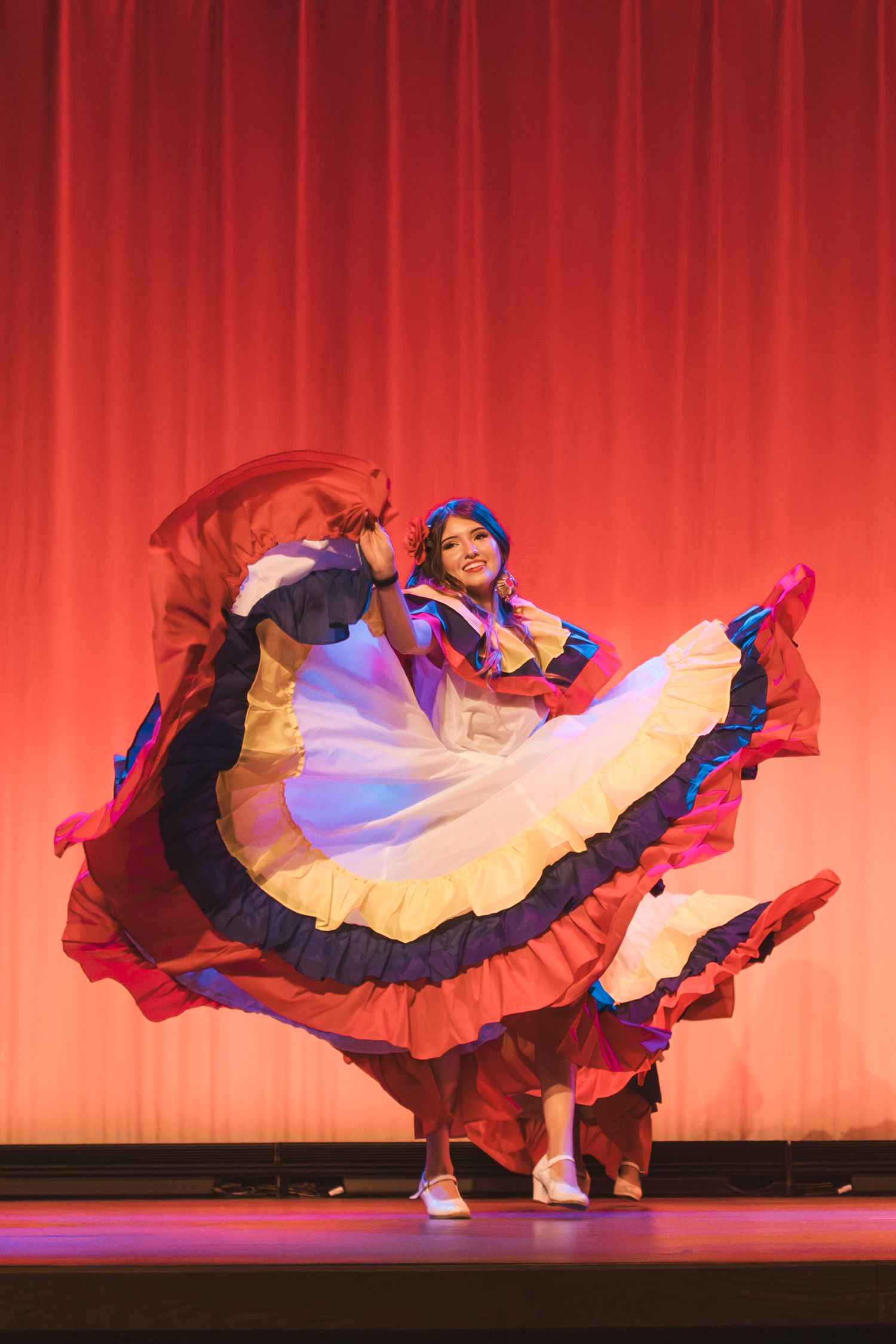 Performer in colorful traditional Latin dress twirls on stage with flowing skirt under red curtain backdrop.