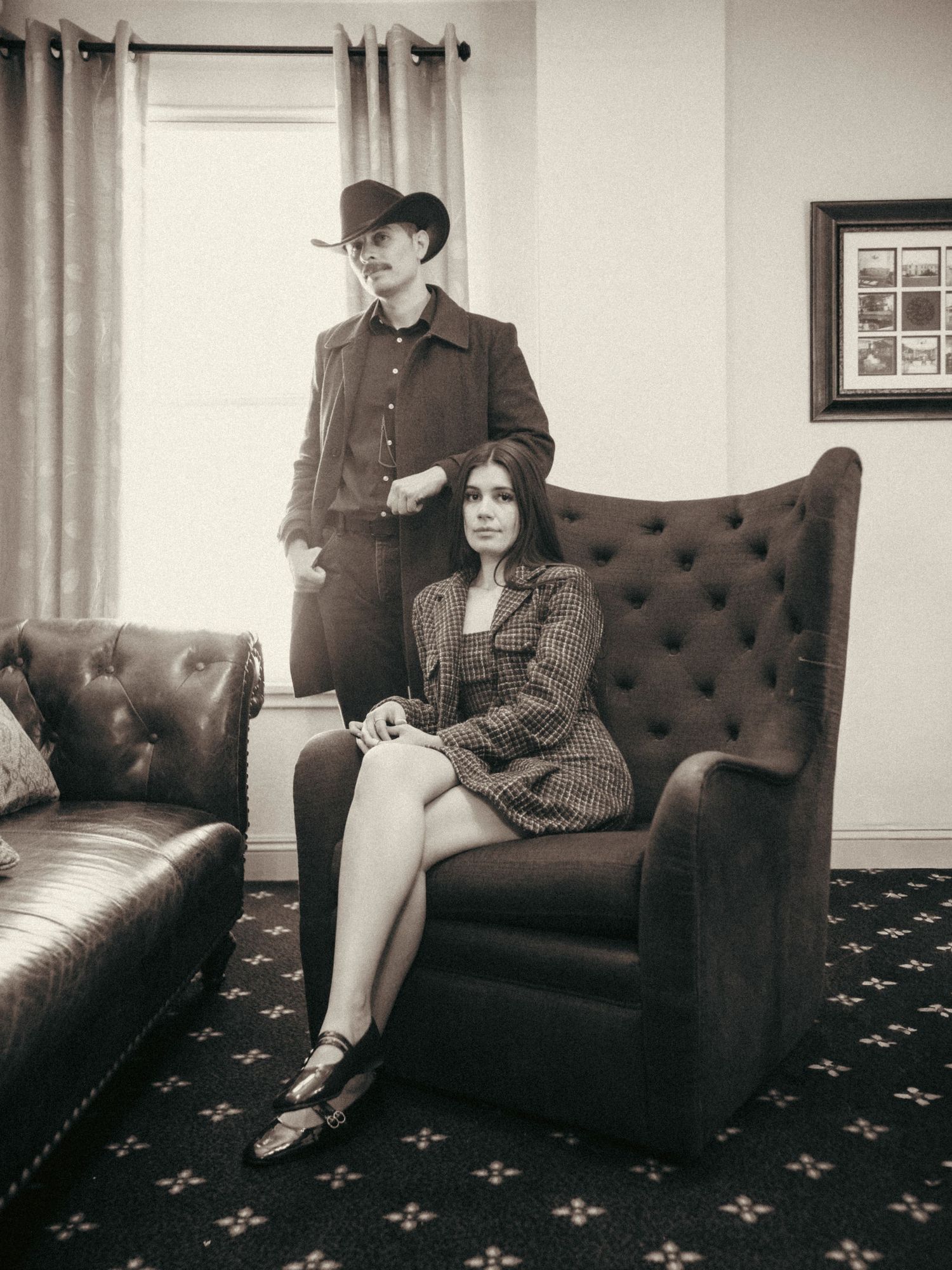 Vintage sepia photo of two people in retro outfits, woman seated in tufted chair, man standing in cowboy hat.