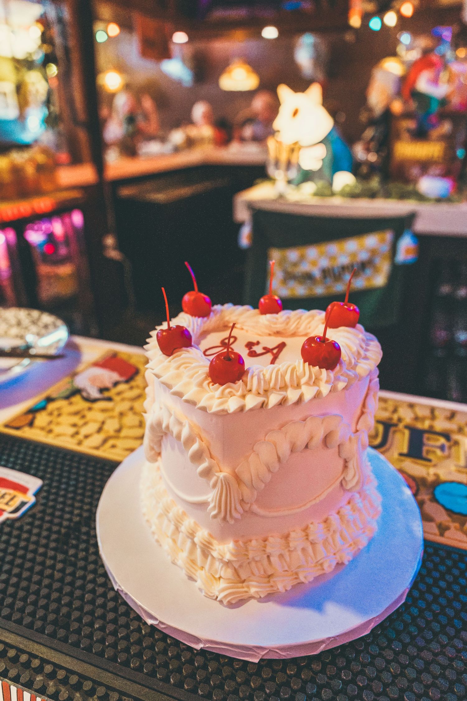 Two-tier white frosted birthday cake topped with maraschino cherries, displayed in a vibrant bar setting.