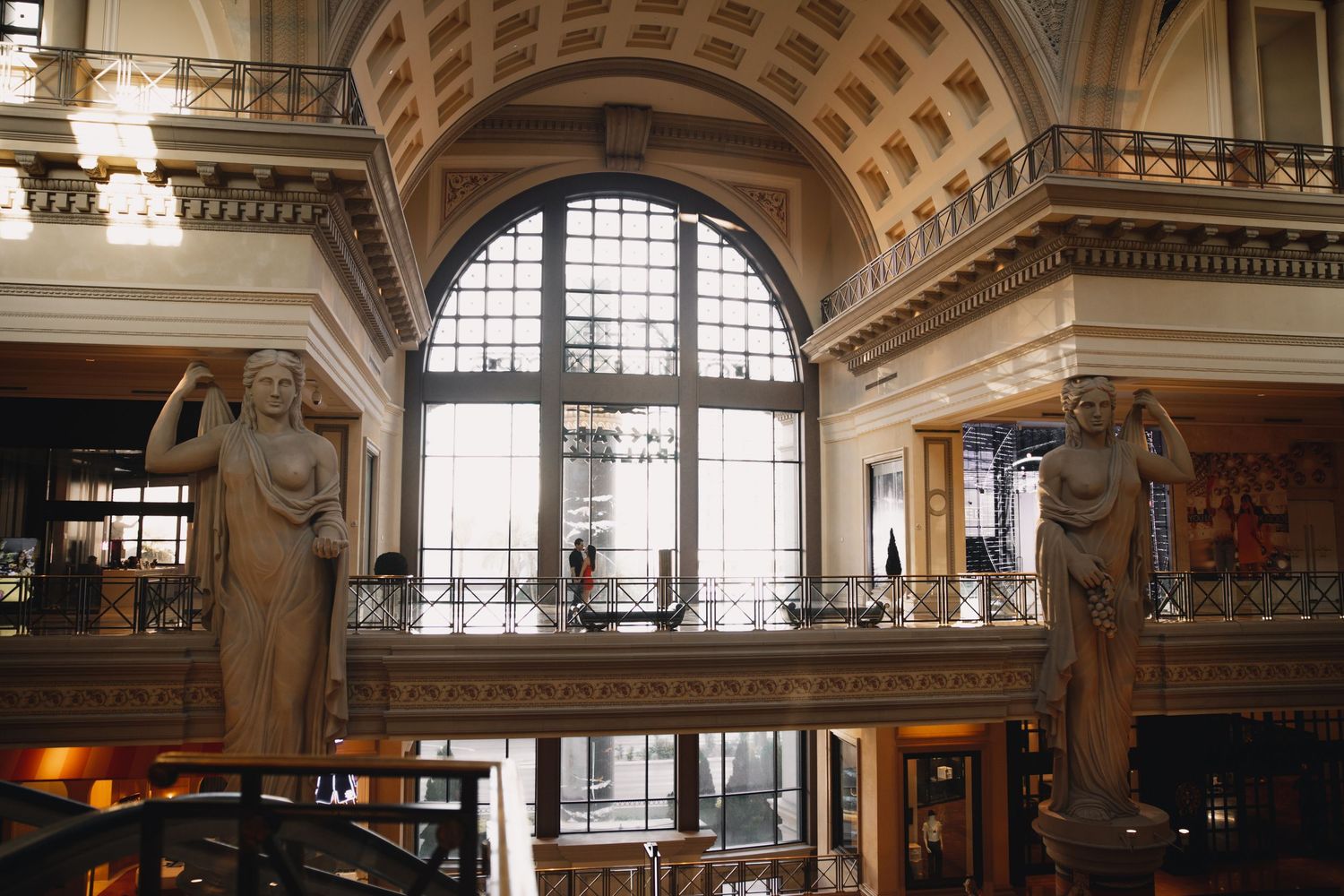 Grand interior of elegant building featuring ornate balcony, arched window, and classical female sculptures on either side.