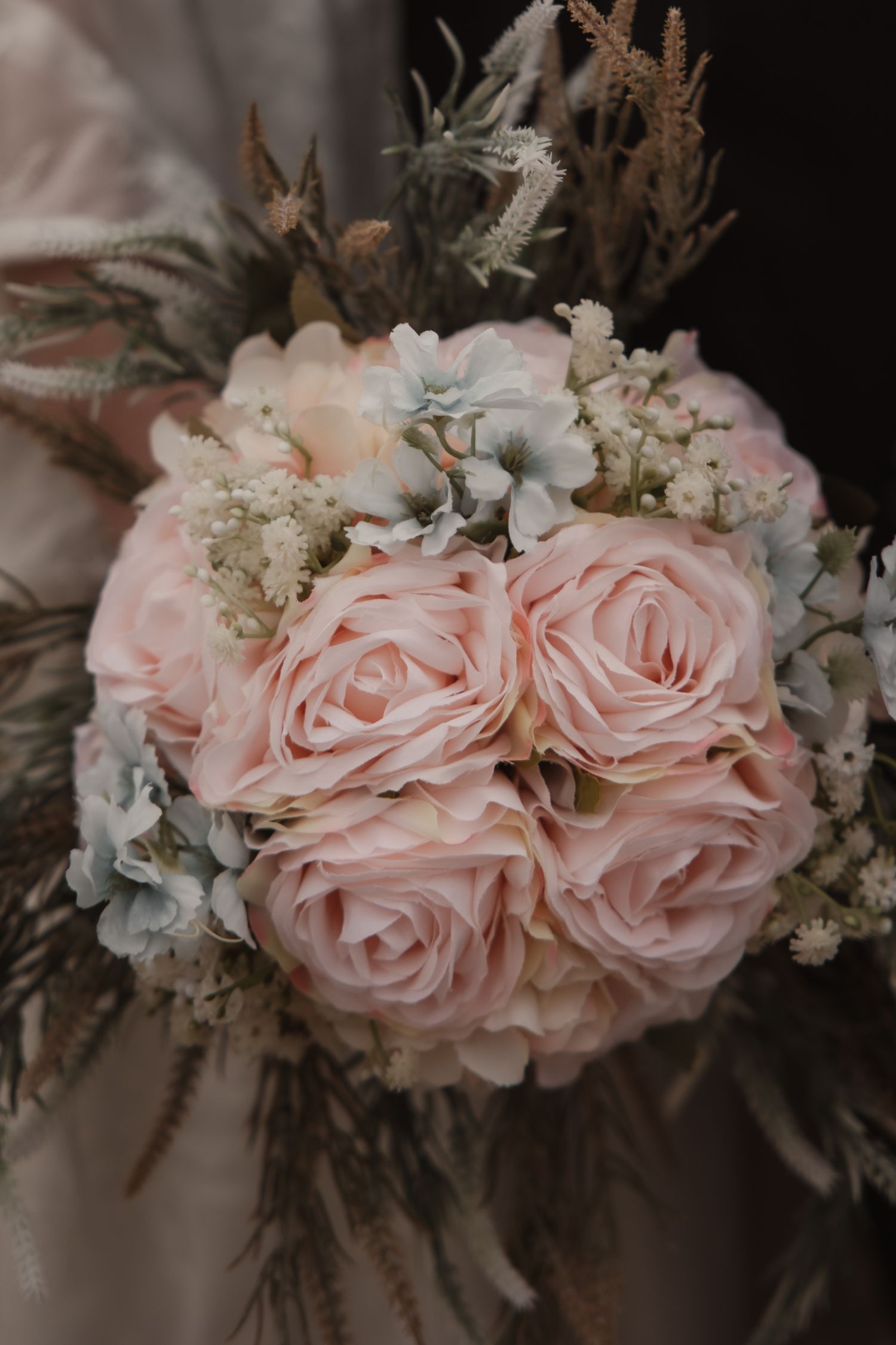Bridal bouquet featuring soft pink roses, blue hydrangeas, and dried grasses held by bride.