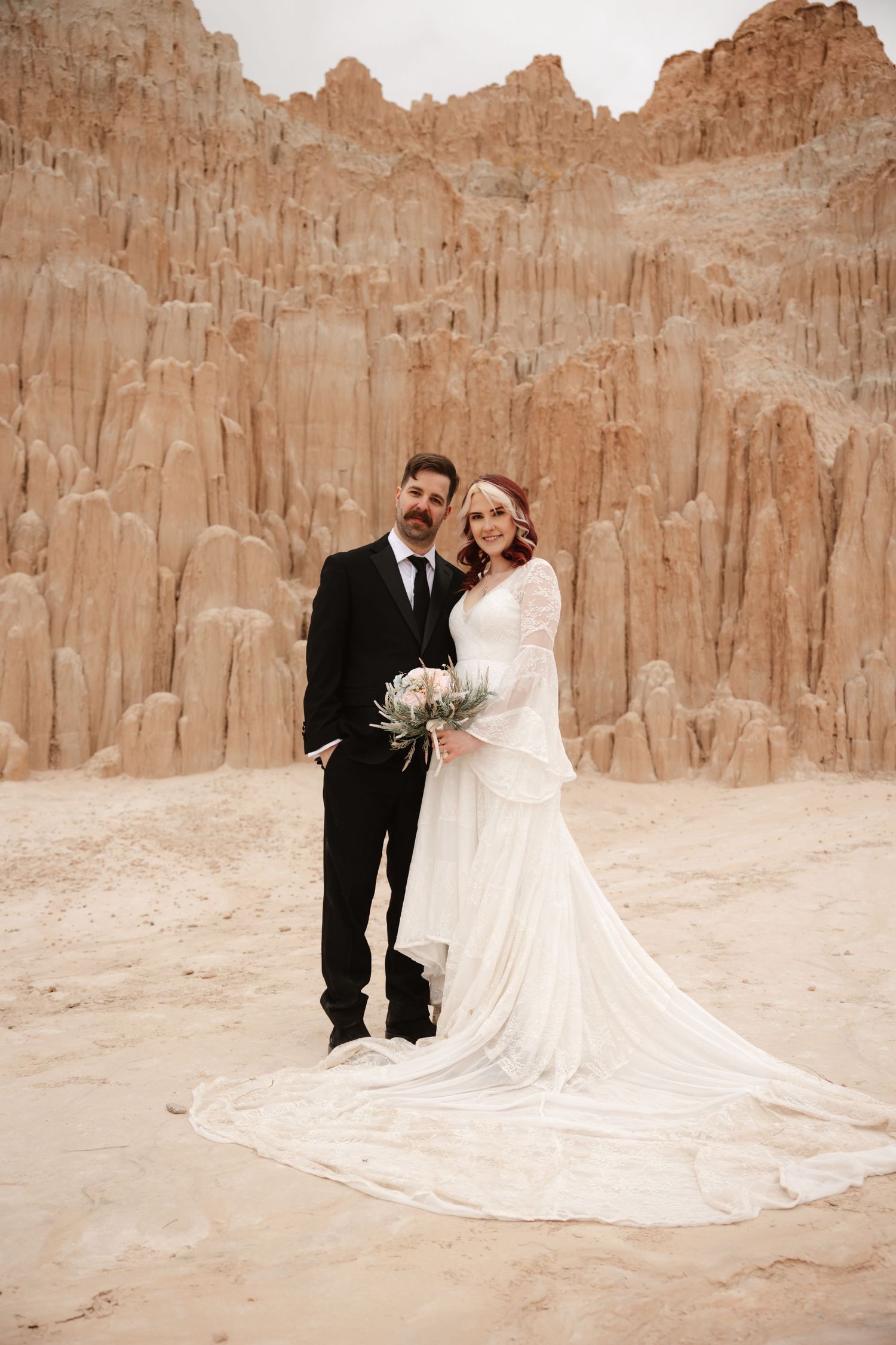 Bride and groom pose together in desert sand surrounded by dramatic eroded sandstone spire formations.