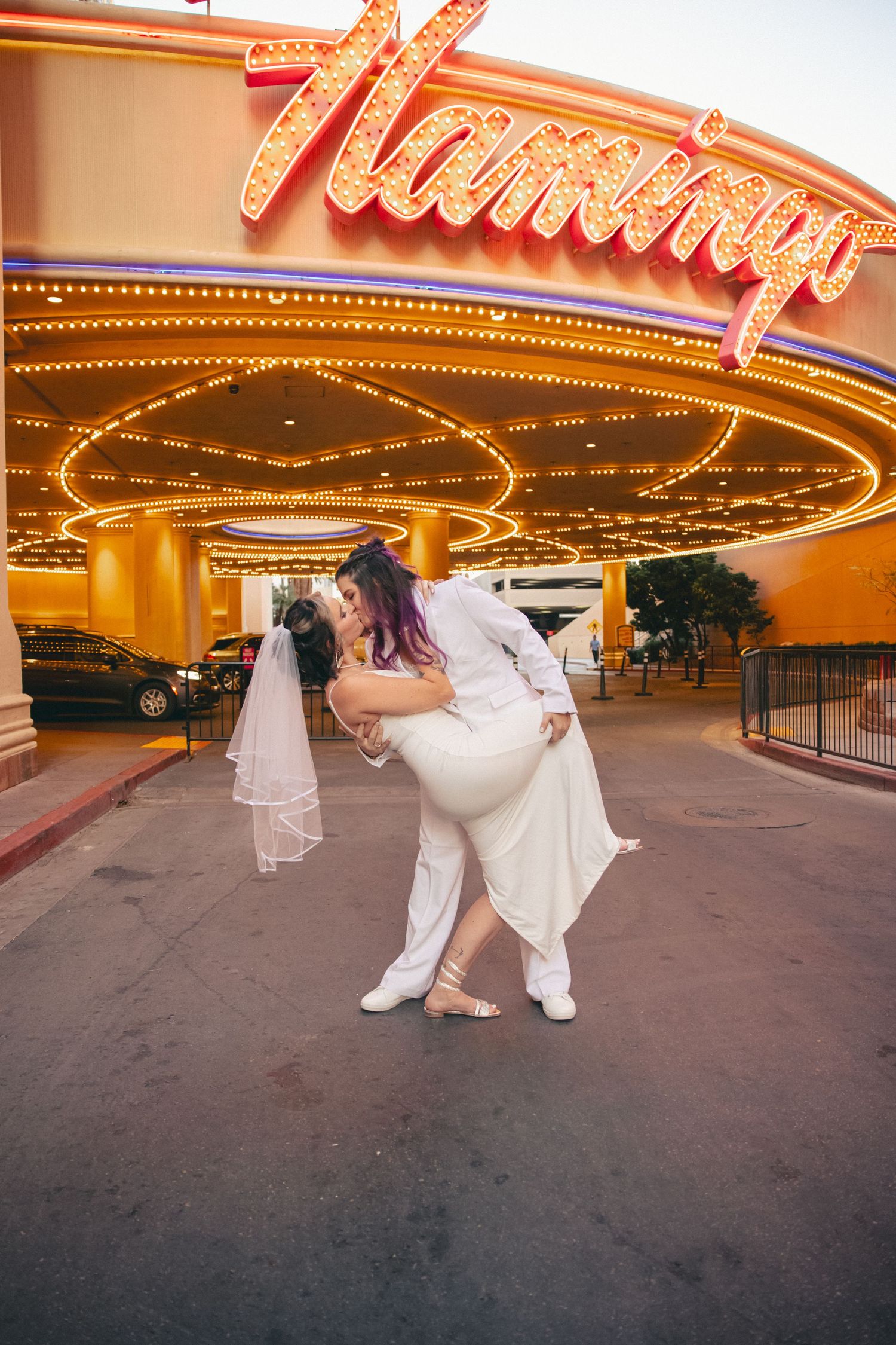 Two brides share a romantic dip kiss in front of the illuminated Flamingo Las Vegas sign.