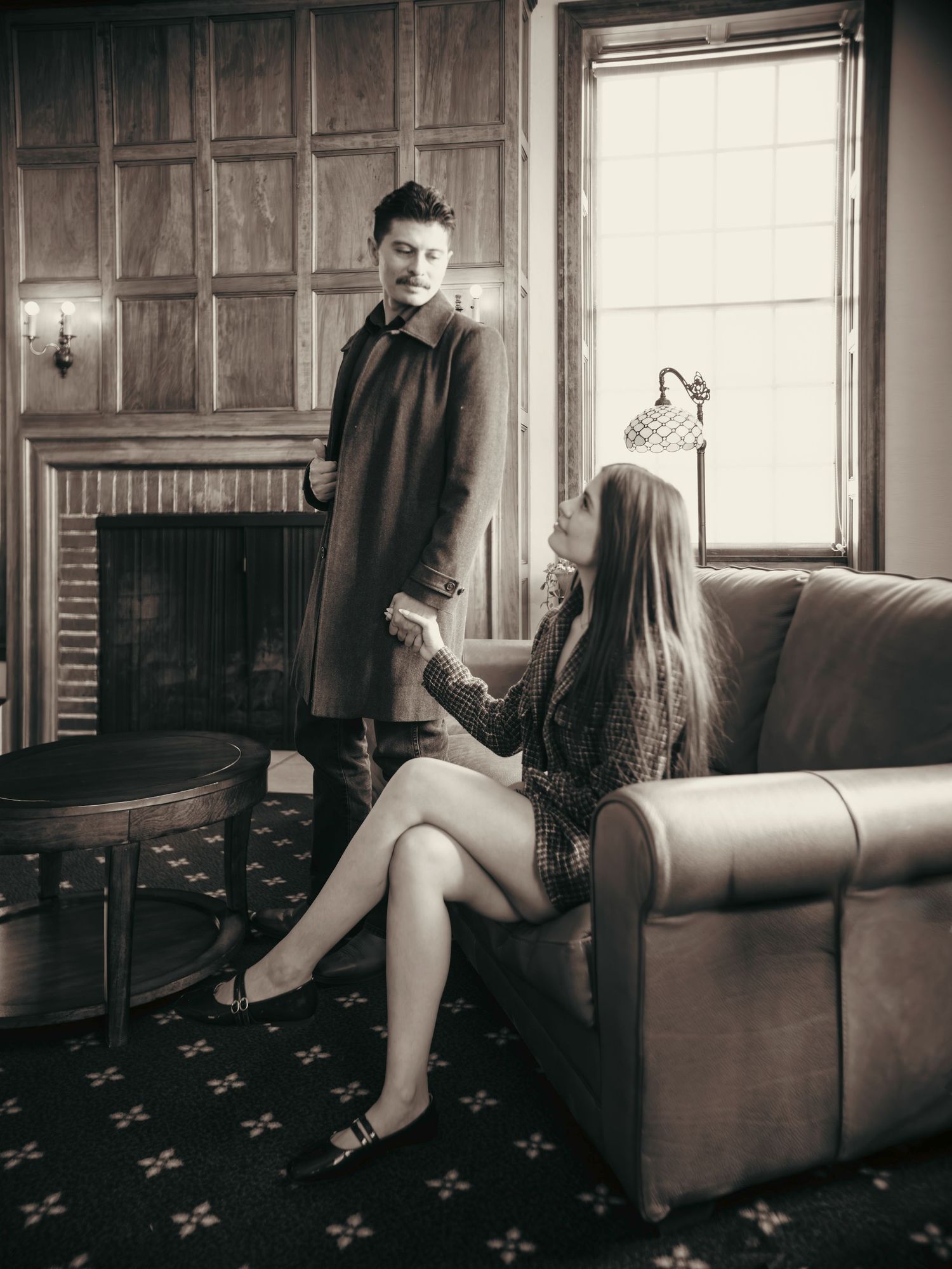 Black and white photo of a couple holding hands, woman seated on leather chair, man standing by fireplace in coat.