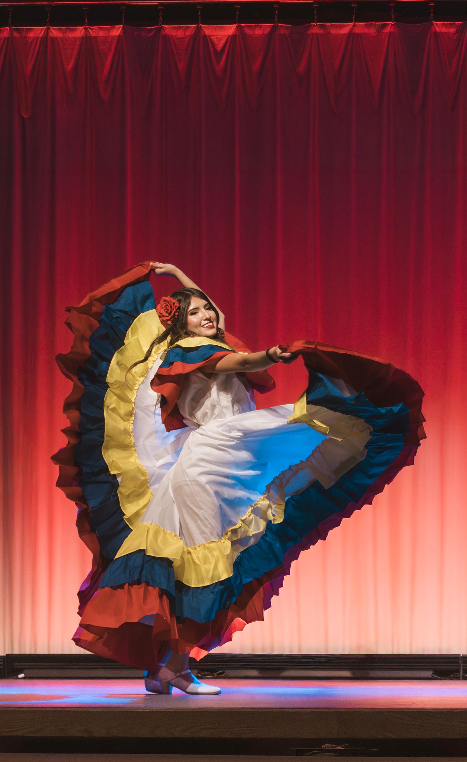 Vibrant folkloric dancer in colorful traditional dress twirls on stage with red curtain backdrop.