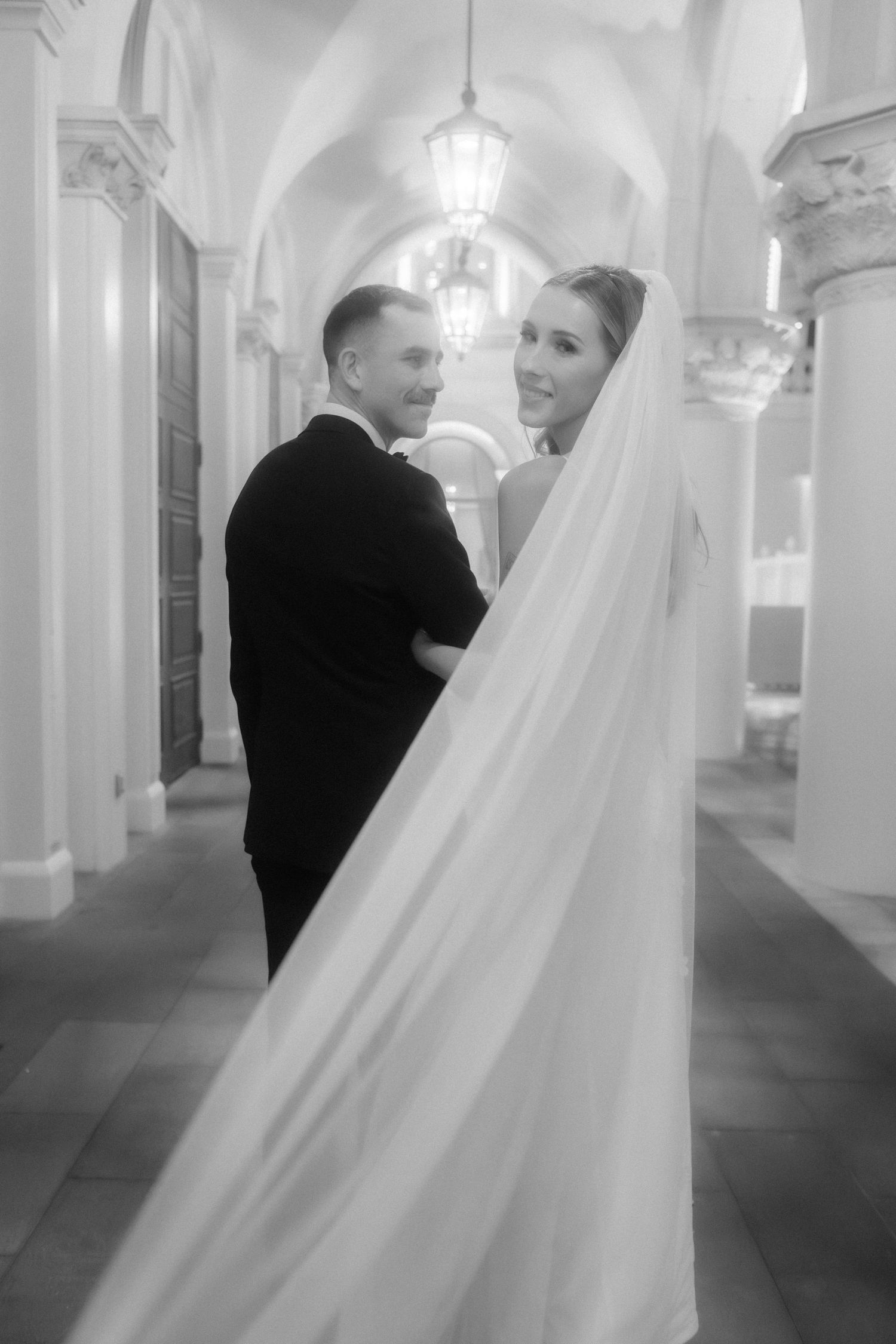 Black and white wedding photo of couple in ornate corridor with veil flowing.
