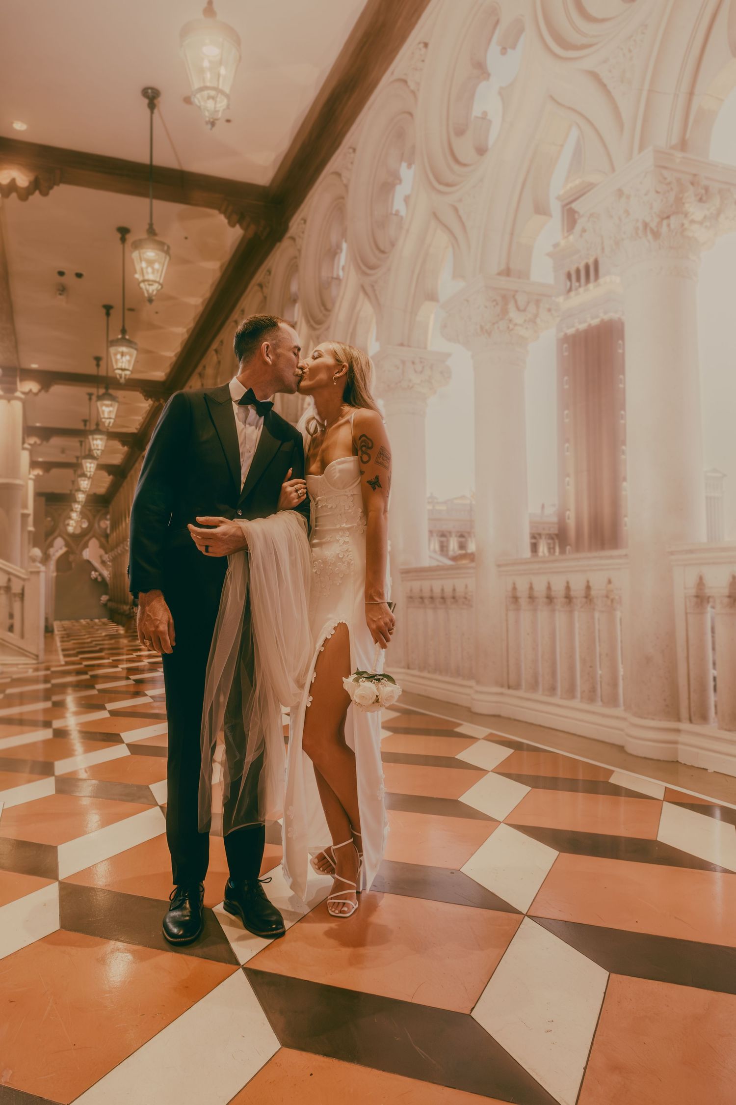 Bride and groom share passionate kiss in grand ornate hallway with warm golden lighting.