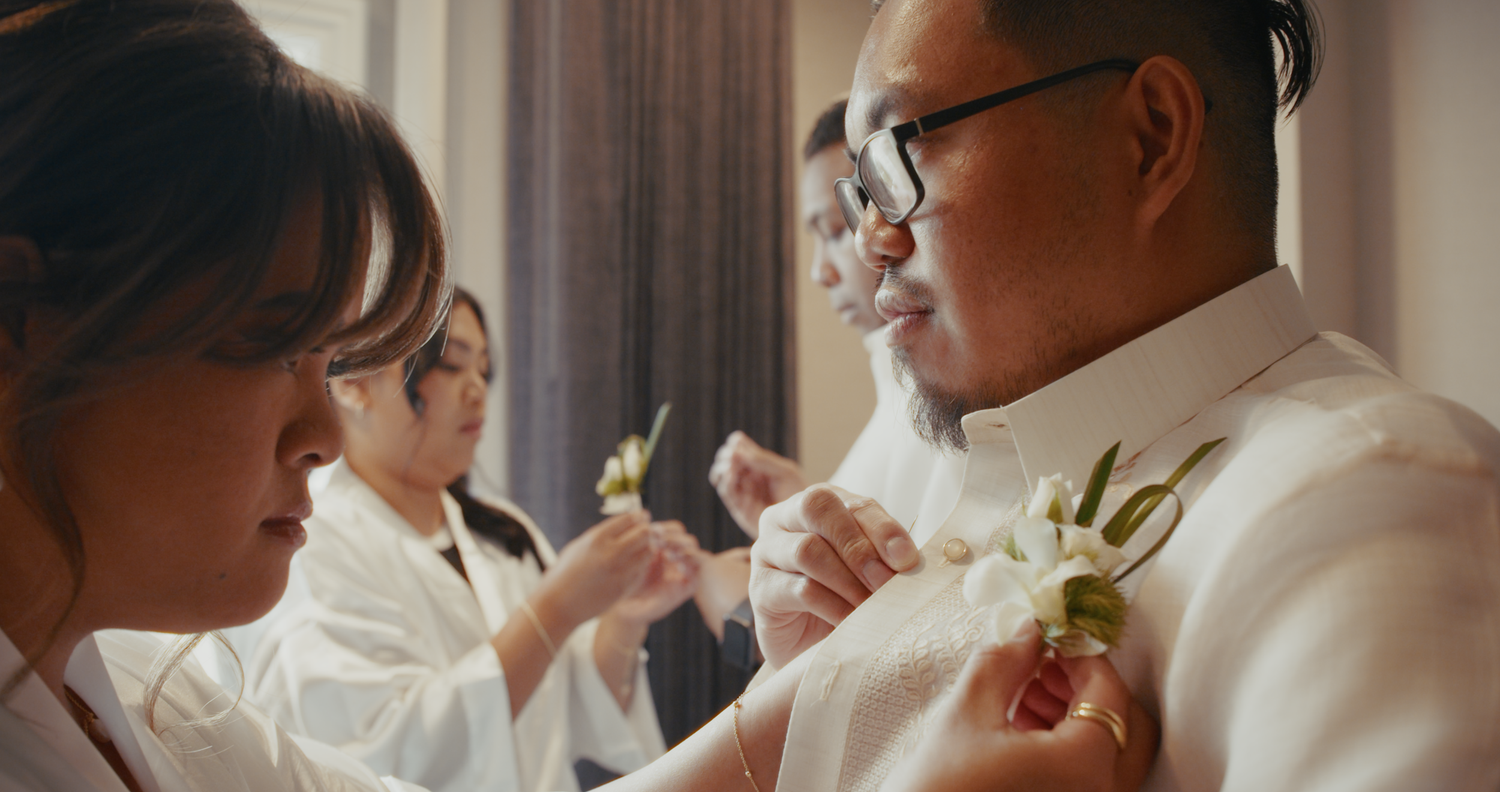 A woman pins a white boutonniere onto a groom's white barong shirt before a wedding ceremony.