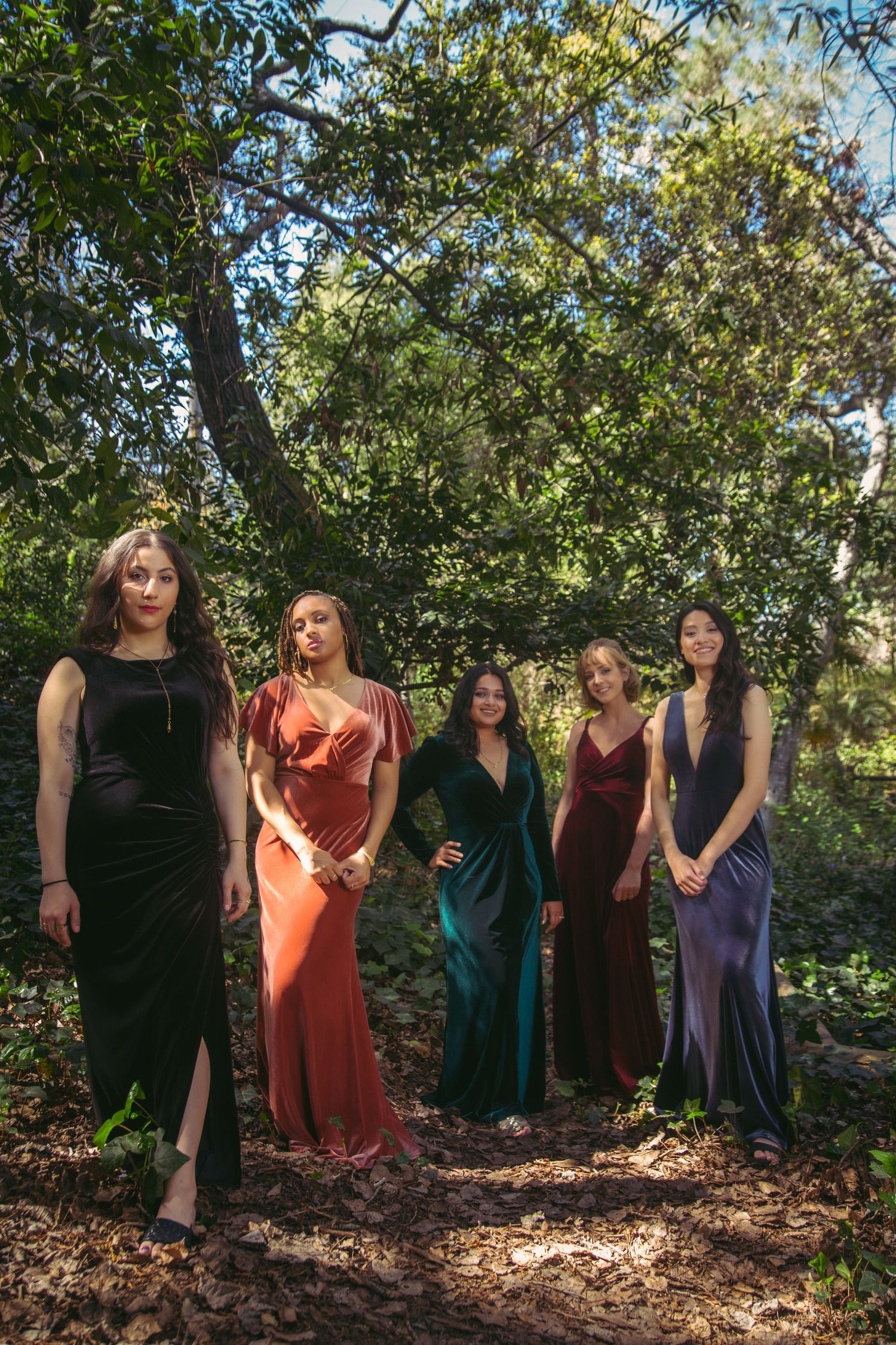 Five women in elegant velvet gowns pose dramatically under a large tree in a forest setting.