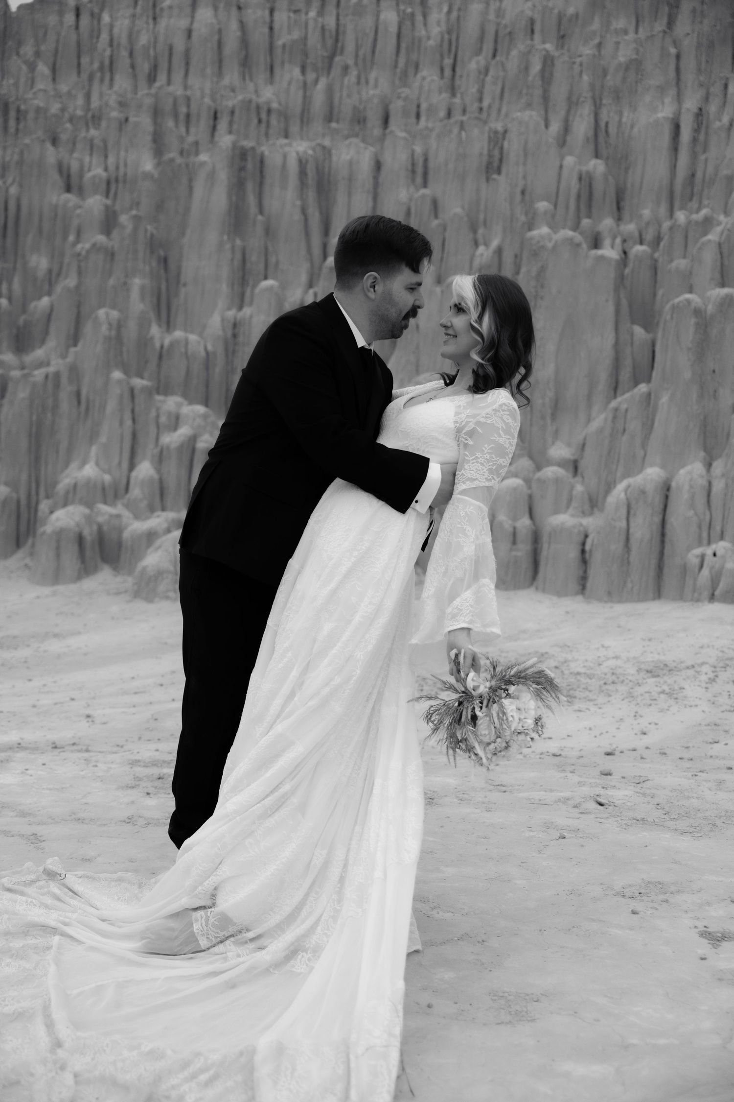 Black and white photo of groom dipping bride before dramatic sandstone formations during desert wedding shoot.