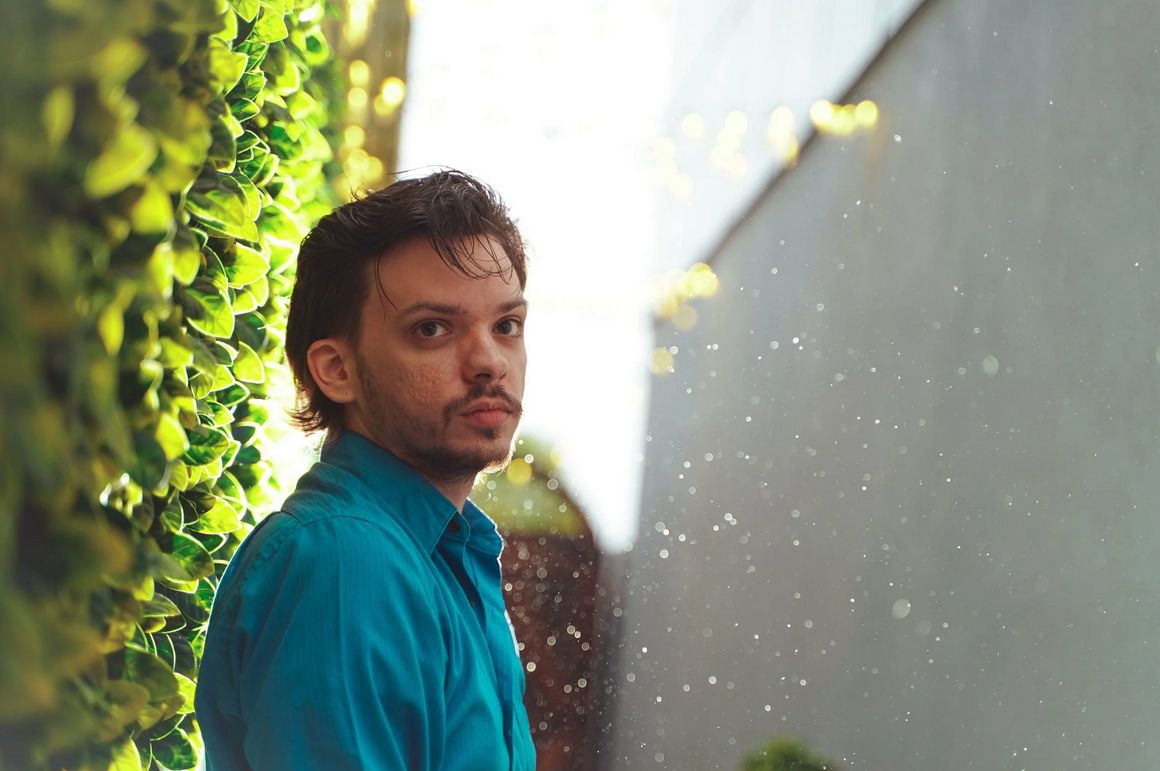 Man in teal shirt leaning against wall beside lush green ivy outdoors.
