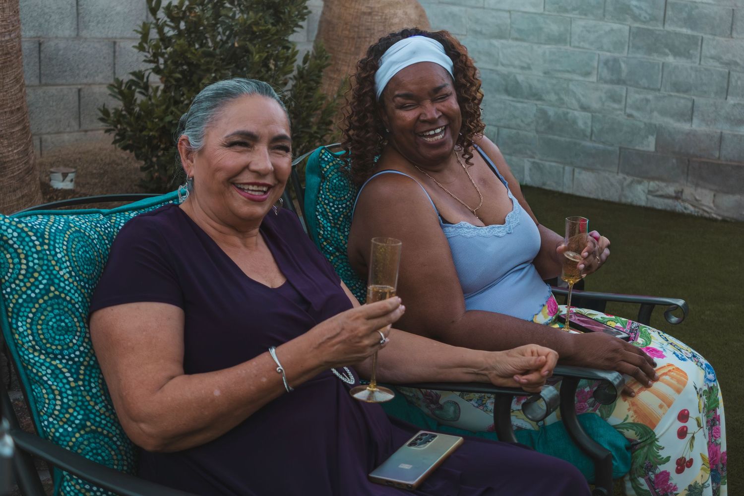 Two women smiling and toasting with champagne glasses while sitting in chairs outdoors at a backyard gathering.