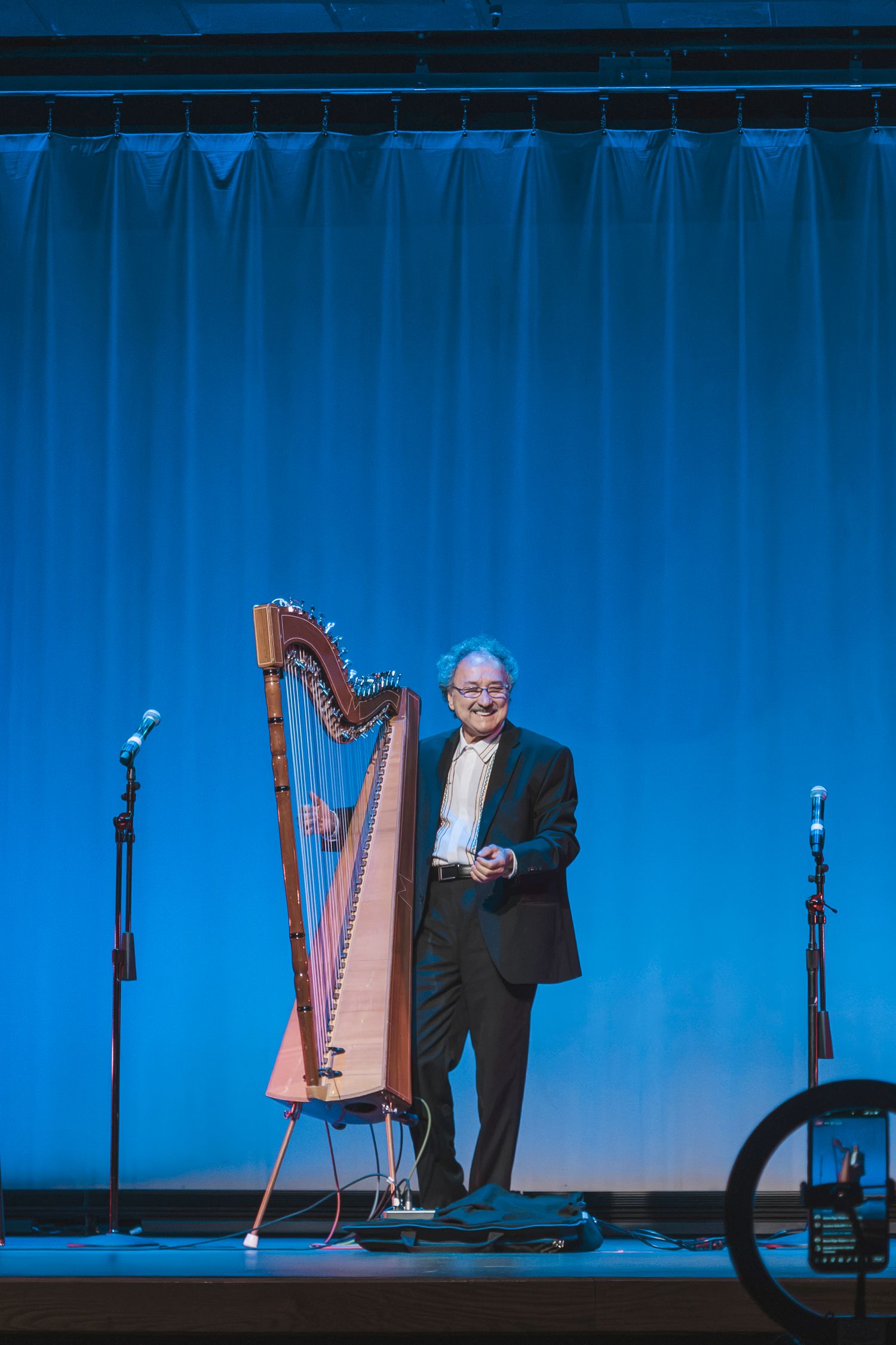 Man in black suit stands smiling on stage beside a wooden harp under blue lighting.