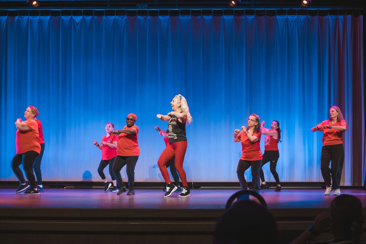 Group of dancers in red outfits performing energetic hip-hop routine on a blue-curtained stage.
