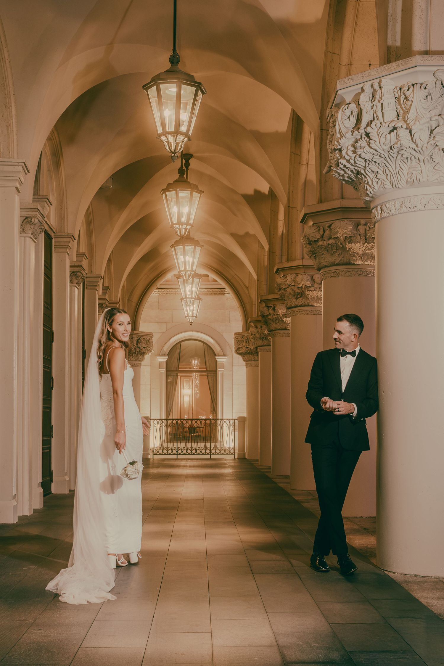 Bride and groom stand apart in an elegant arched corridor with hanging lanterns during their wedding shoot.