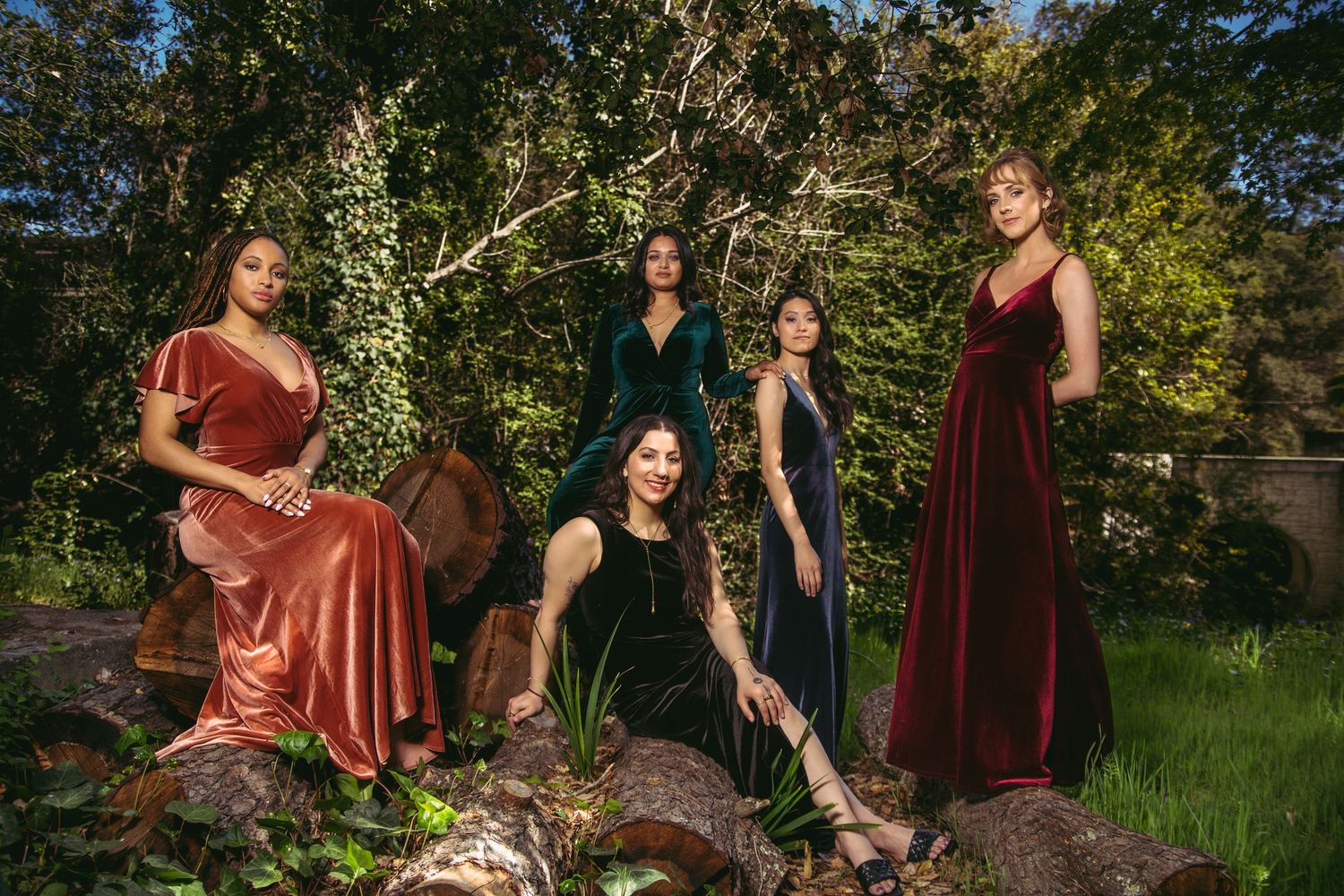 Five women in velvet evening gowns posing on logs in a lush garden setting.