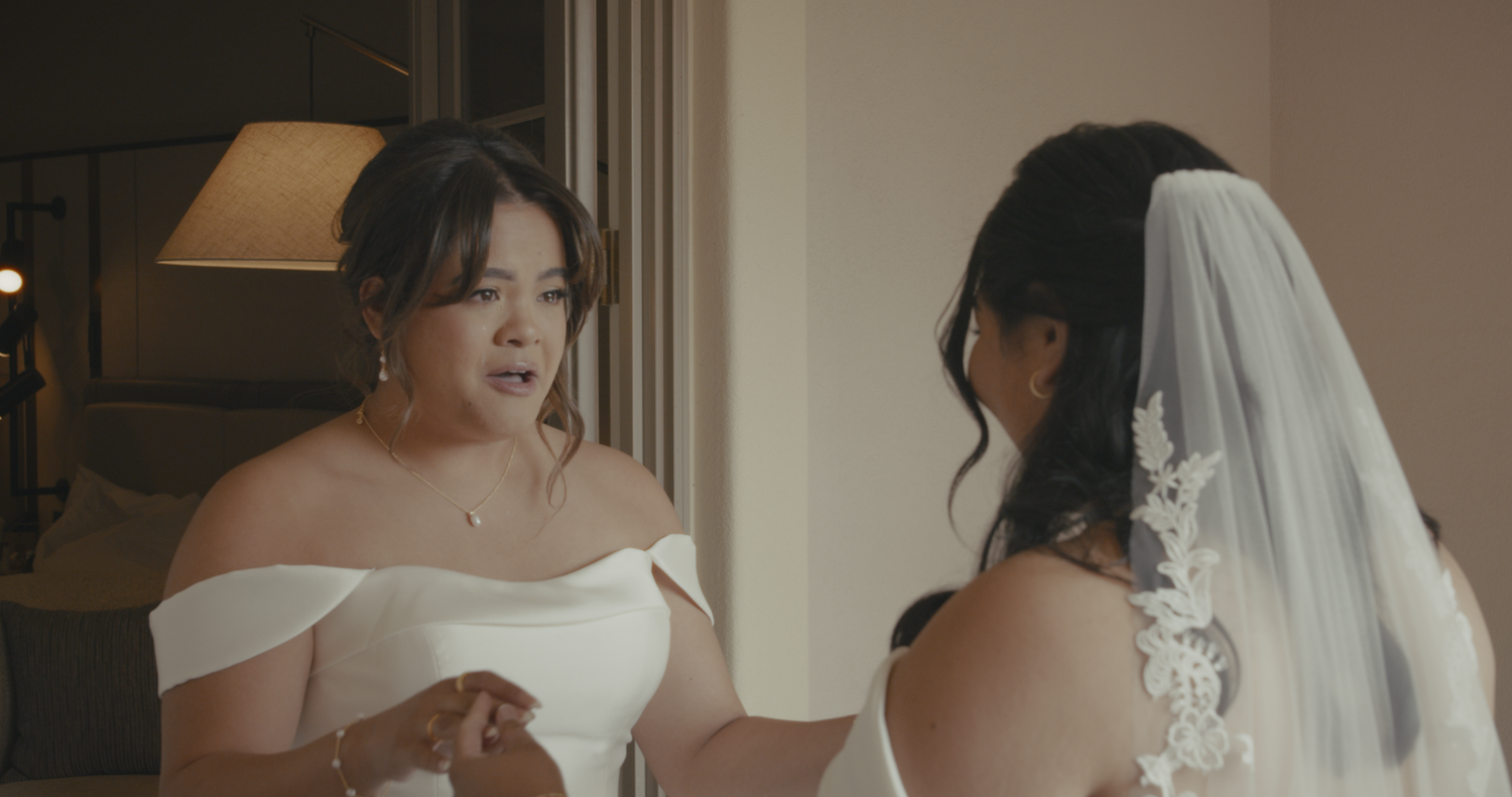 Two women in white off-shoulder bridal gowns share an emotional moment indoors.