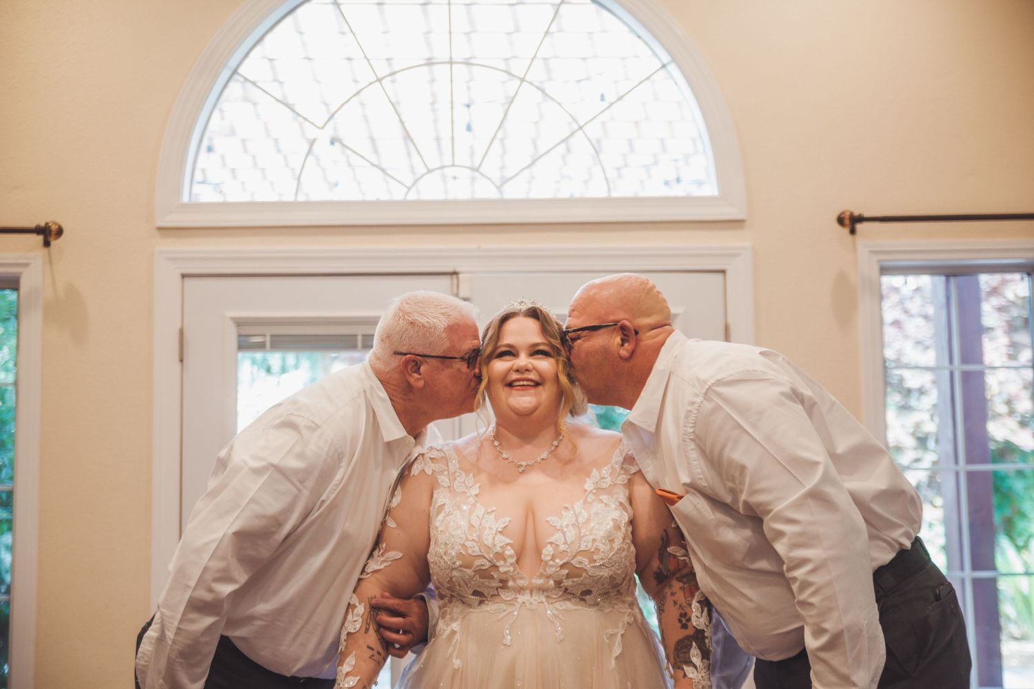 Bride in lace wedding gown smiles as two men in white shirts kiss her cheeks near elegant arched window.