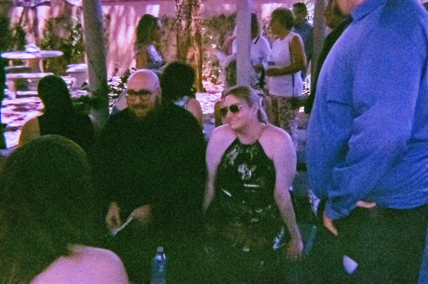 A bald man and a blonde woman in a black halter dress socialize at an outdoor evening party with purple lighting.