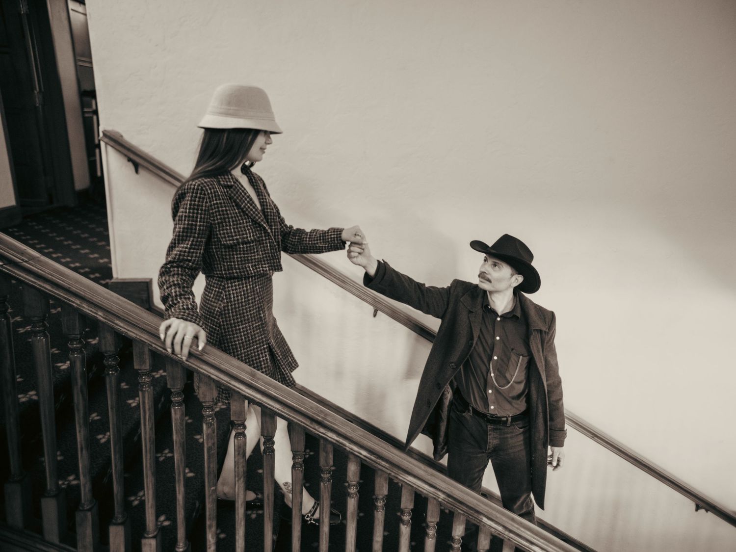 Two people in vintage Western attire exchange a handshake on a staircase in a sepia-toned photo.