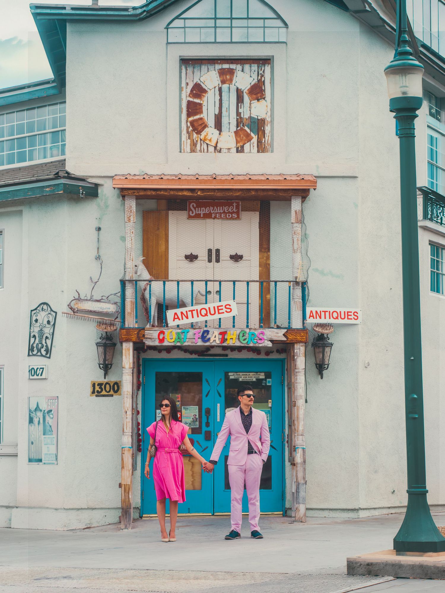 Couple in pink outfits holding hands outside a colorful antiques shop with teal doors and a balcony.
