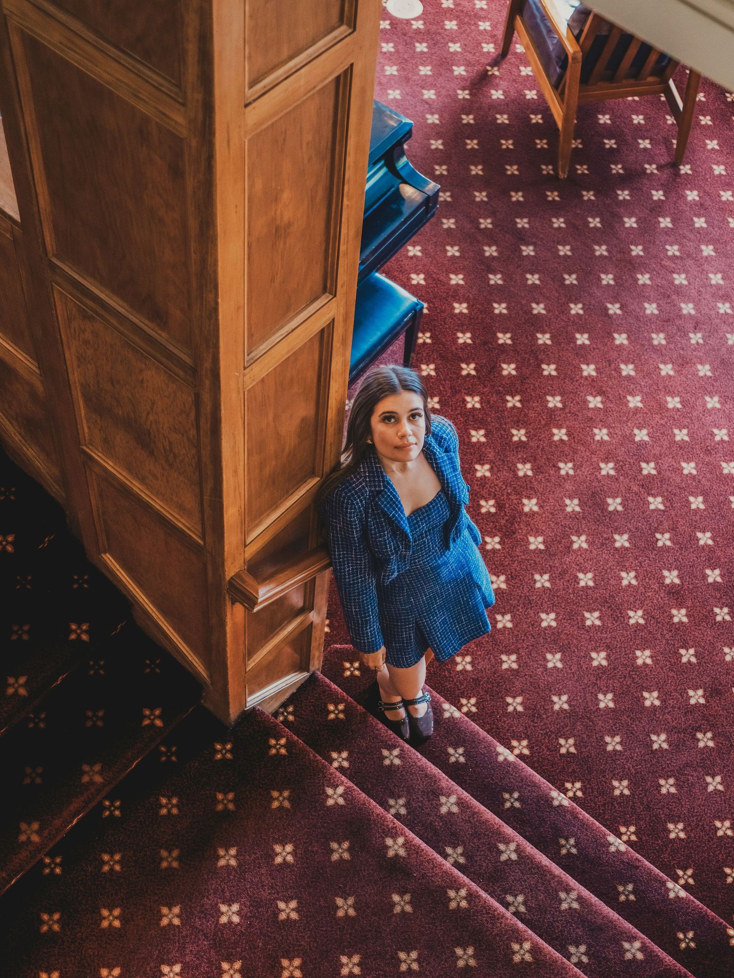 Woman in blue outfit standing at the base of stairs in an elegant venue with patterned red carpet.