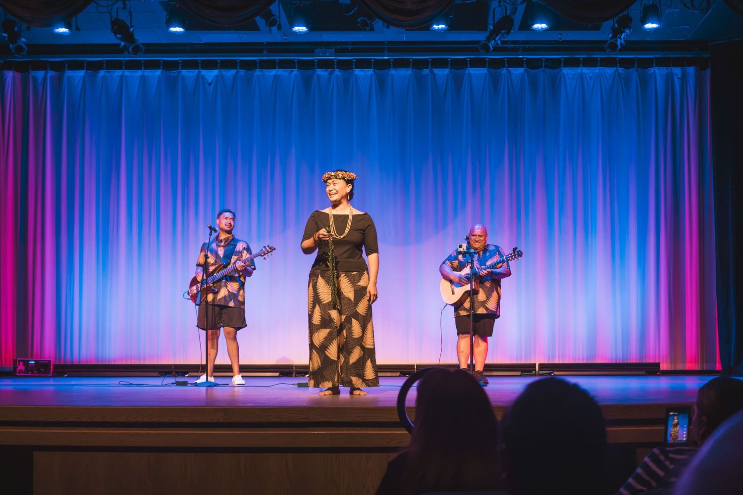 Three performers on stage, two playing ukuleles and one singing, in front of a blue curtain backdrop.