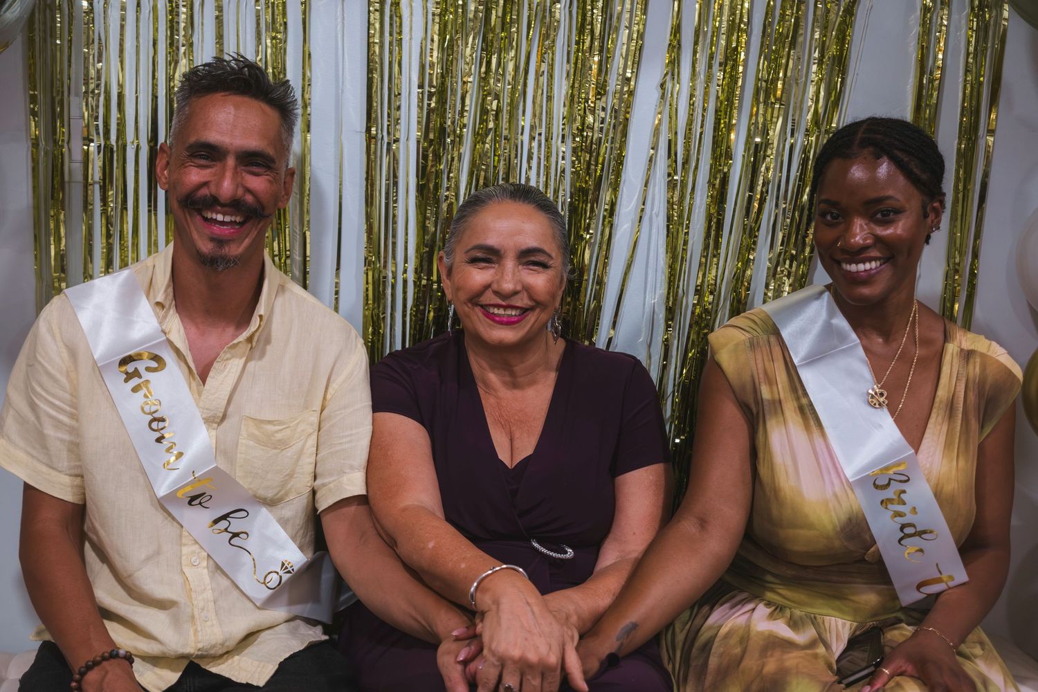 Three smiling people pose together in front of a gold tinsel backdrop, two wearing celebratory sashes at a festive event.