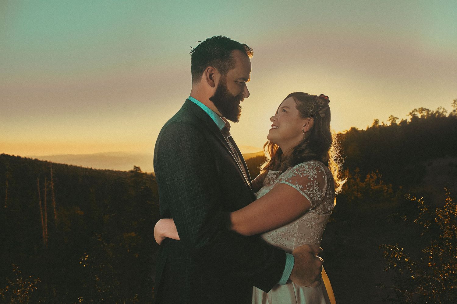 Couple embracing at sunset in a vineyard, groom in suit and bride in white dress, golden sky backdrop.