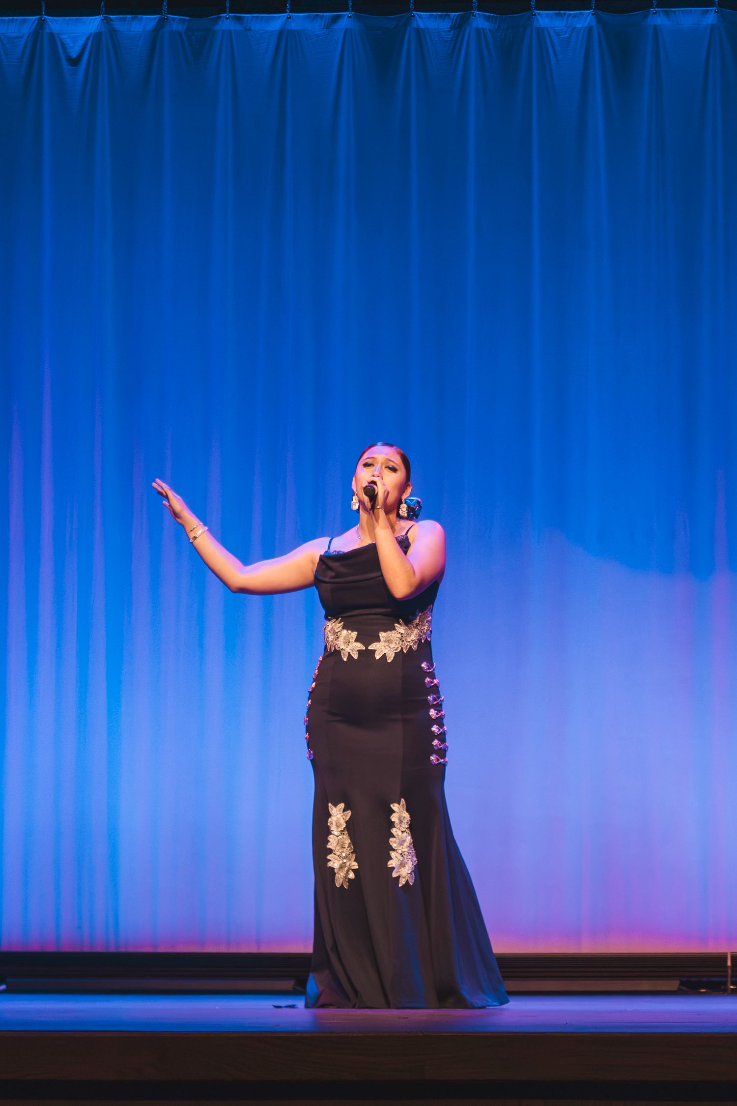 Woman in black gown with gold lace appliques singing on stage with blue curtain backdrop.
