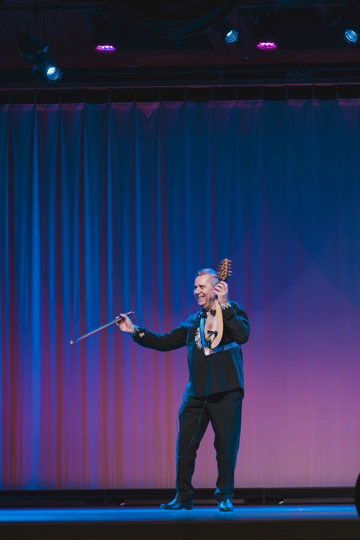 Performer on stage in black suit holding a baton and instrument under blue and pink stage lights.