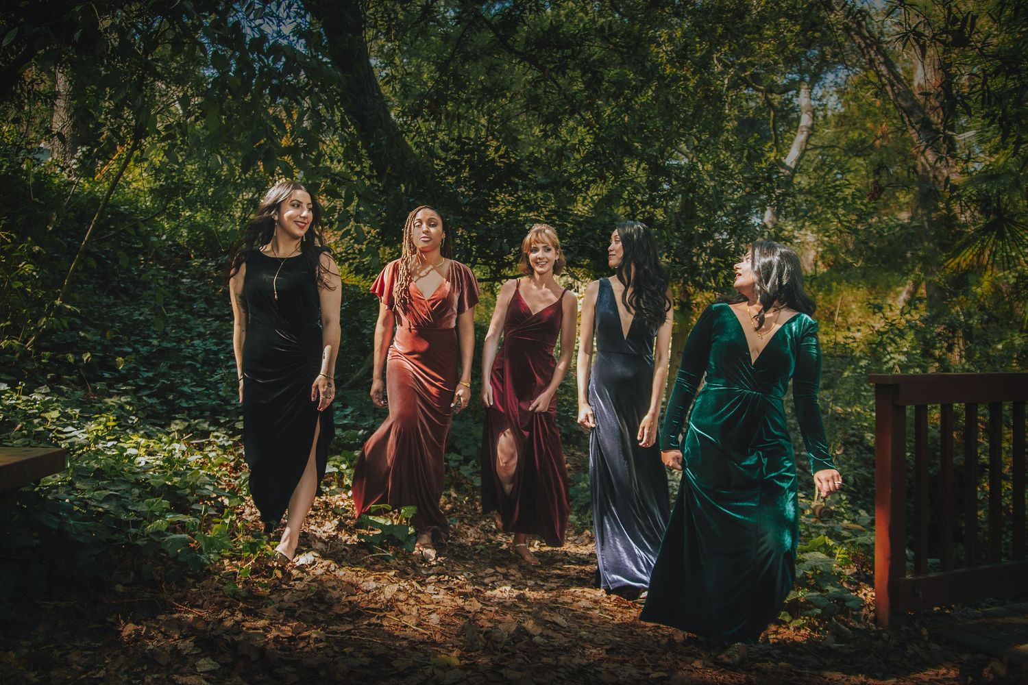Five women in elegant colorful gowns walking through a lush green forest setting.