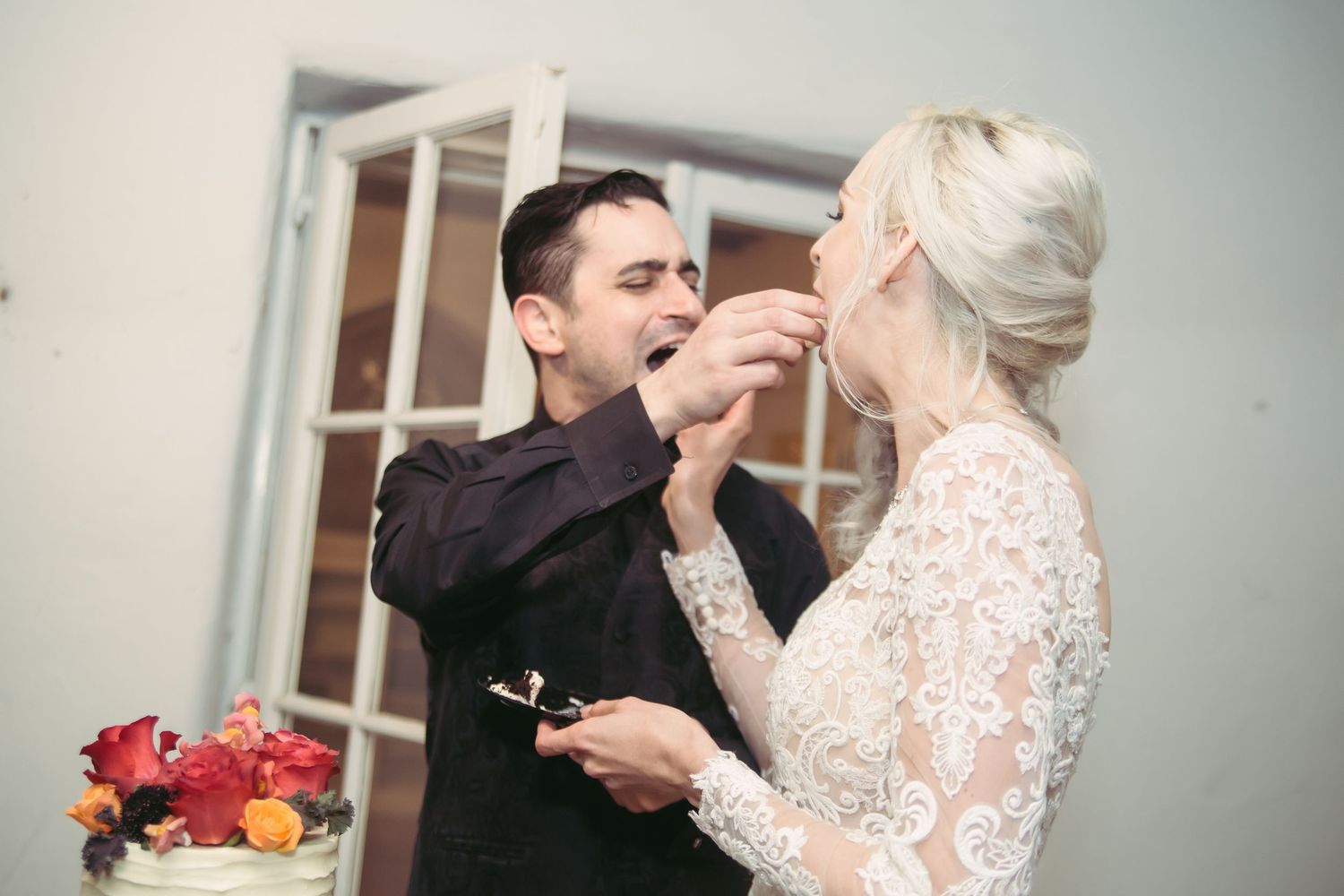 A bride in a lace wedding dress feeds a bite of wedding cake to her groom at their wedding reception.