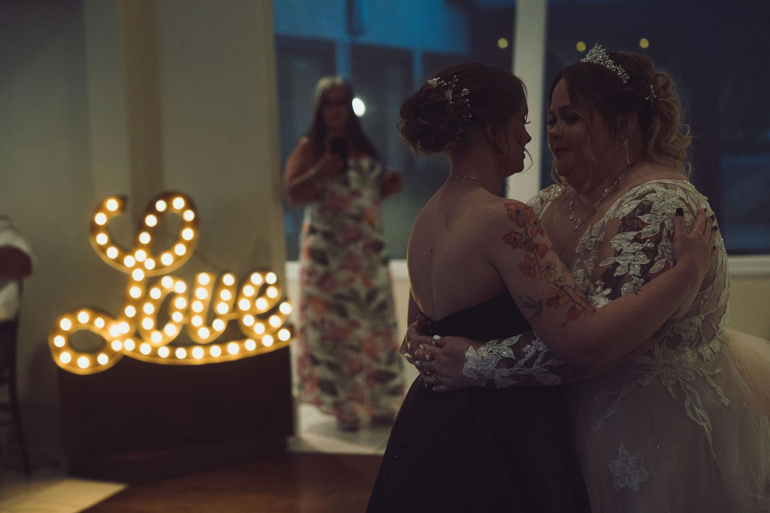 Two women share a first dance near a marquee Love sign at a wedding reception.