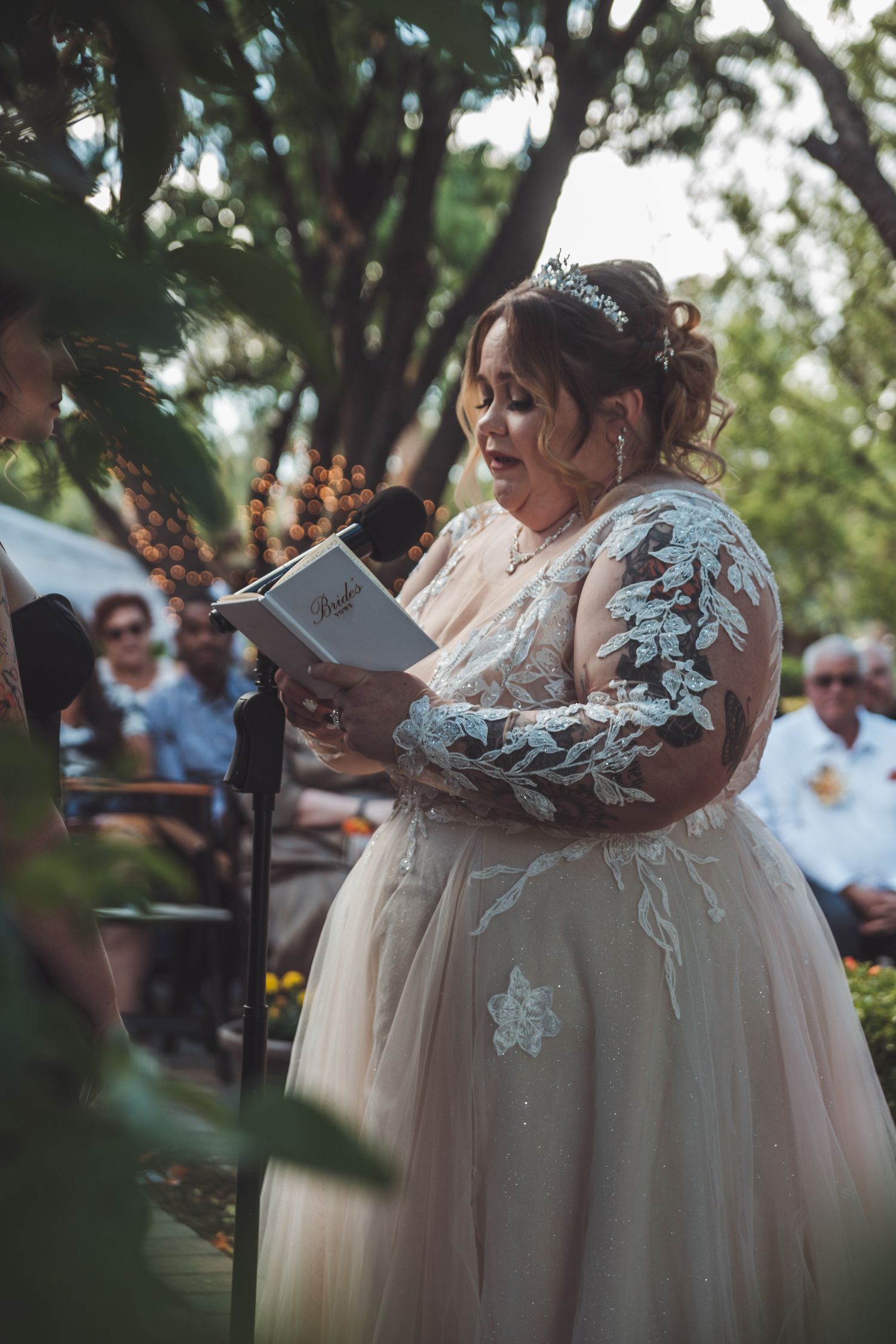 Plus-size bride in lace wedding gown and tiara reads vows from book at outdoor wedding ceremony.