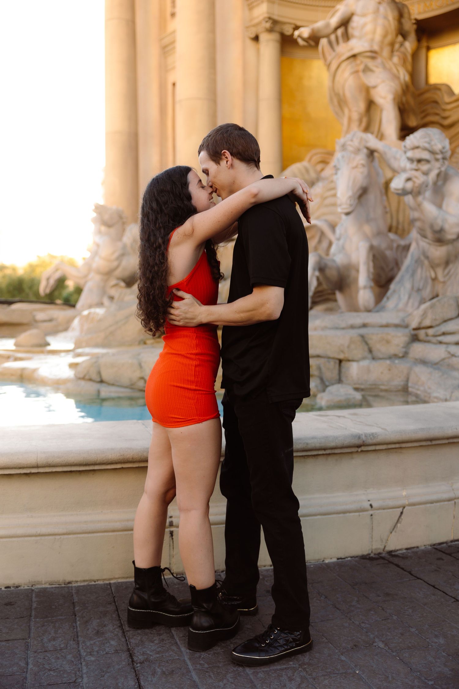 Couple kissing and embracing in front of classical sculpture fountain at golden hour.